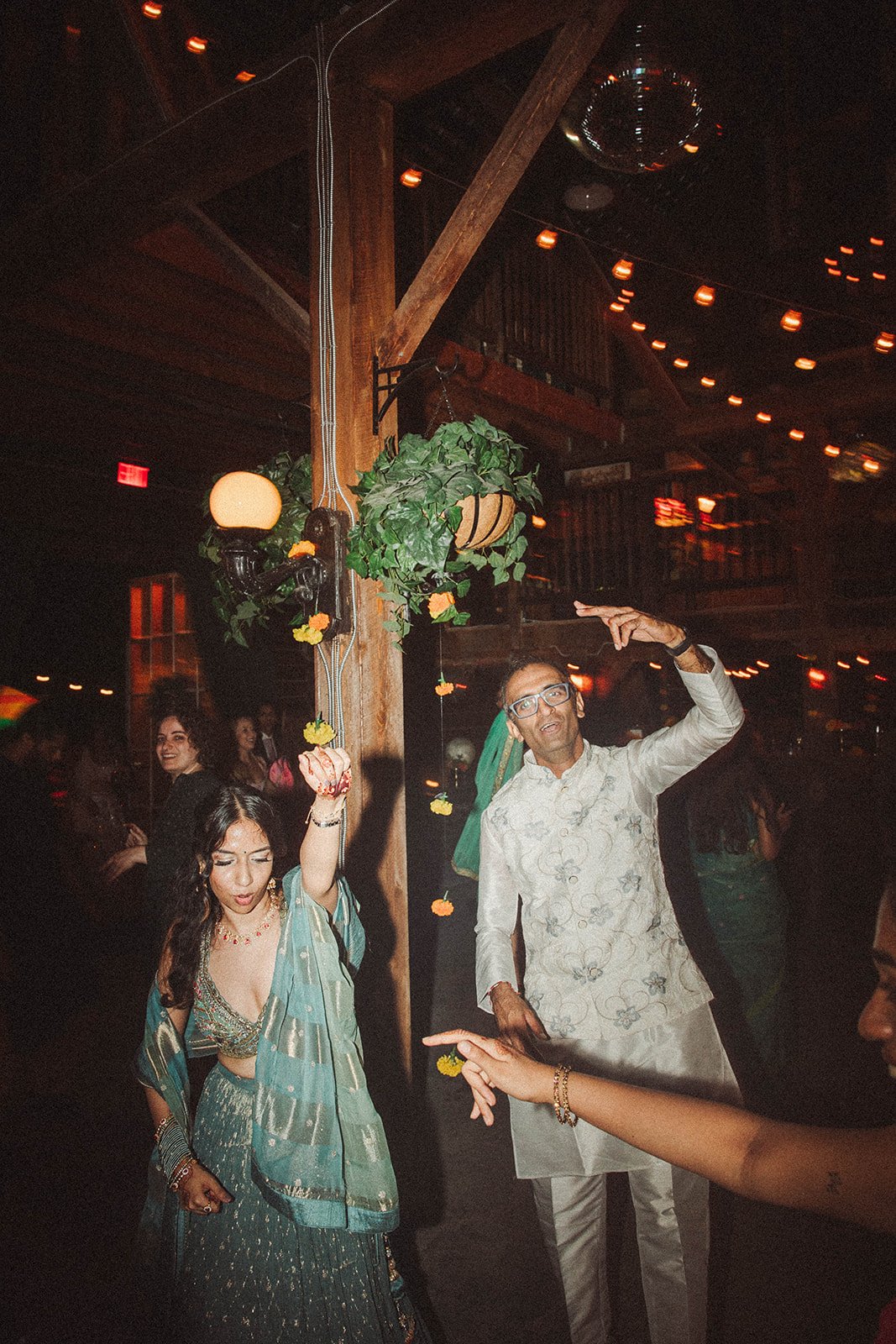 People dancing and celebrating at a party with wooden decor, hanging plants, and string lights.
