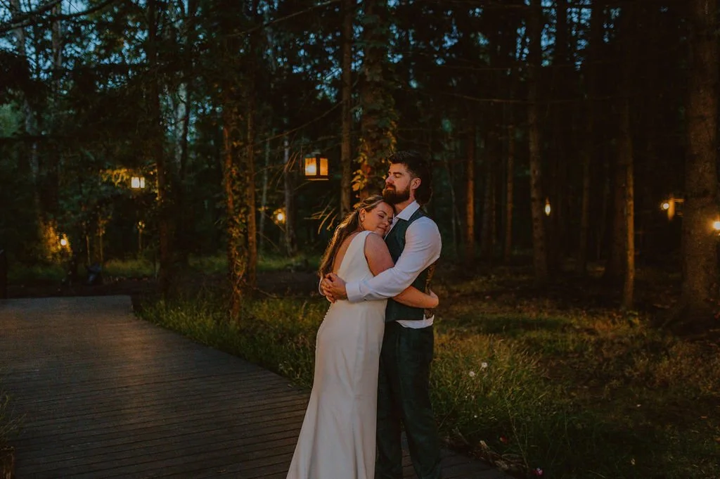 A couple embracing on a wooden path in a wooded area during evening, with lantern lights hanging in the background.