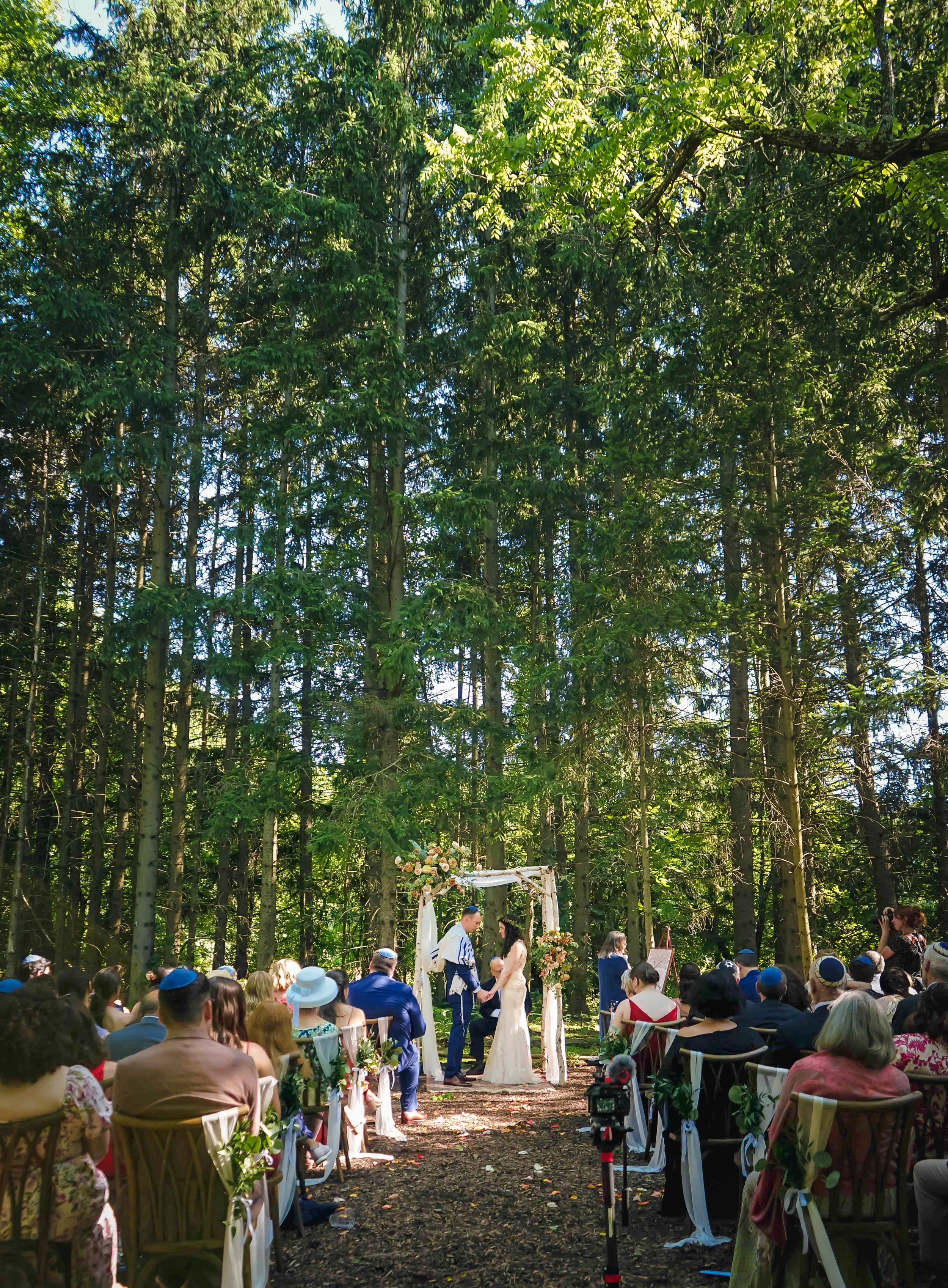 A couple getting married in a forest with an audience seated on wooden chairs, decorated with flowers, under a white floral arch.