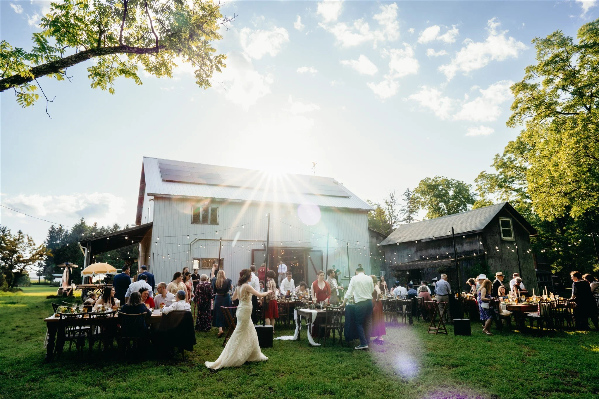 A bride strolls across a lawn filled with rustic dining tables, beneath string lights
