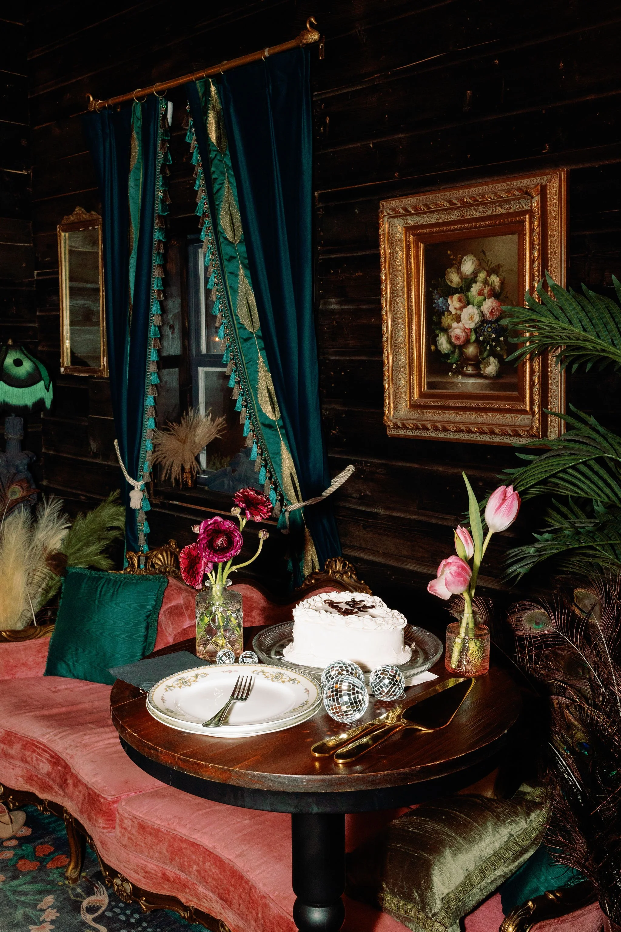 A vintage-inspired room with dark wooden walls, a pink velvet sofa, and gold accents. A round wooden table is set with a white cake, decorative spherical disco balls, and floral arrangements in glass vases. The window is adorned with deep blue, teal,