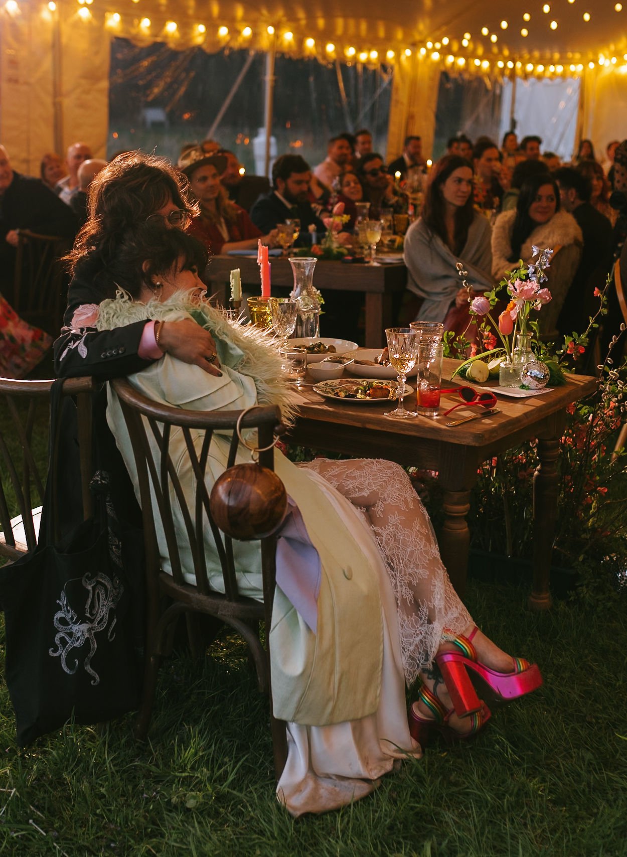 A woman with dark hair and glasses is sitting at a table, hugging a person wrapped in a white blanket, in a tent decorated with string lights. The table is decorated with flowers, candles, and drinks, and there is a large group of people in the backg