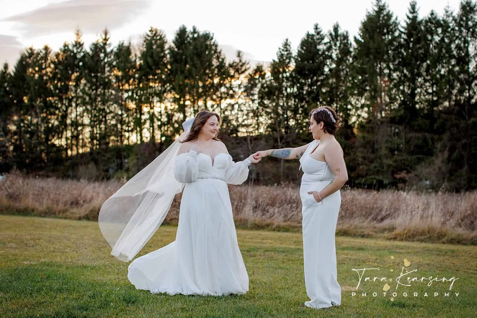 Two women, one in a wedding dress with a veil, and the other in a white jumpsuit, holding hands on a grassy field with trees in the background during sunset.