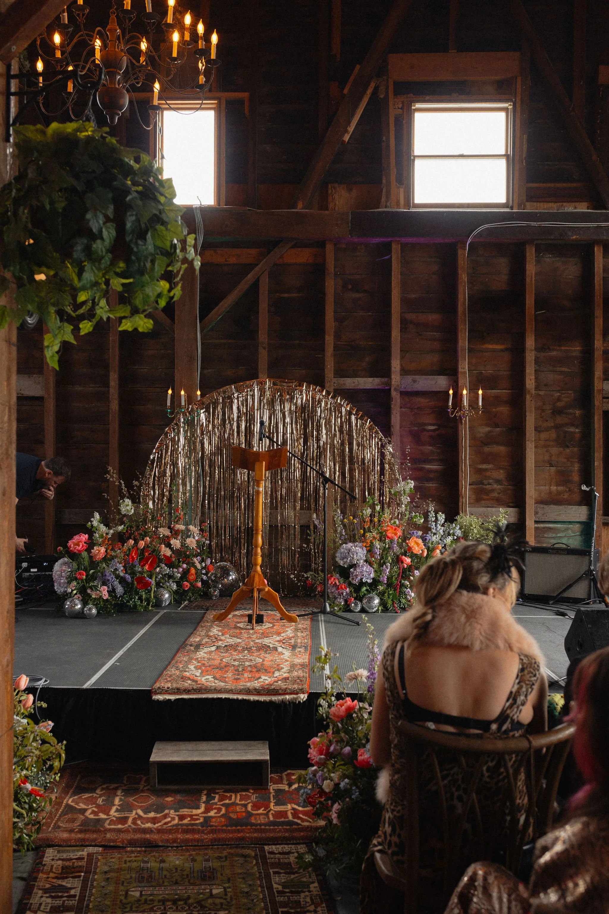 A rustic wooden barn interior set up for a ceremony or event. There is a small stage with flowers and shiny silver orbs, a podium, and microphone. People seated, including a woman in a leopard print dress and fur stole. Vintage rugs on the floor, han