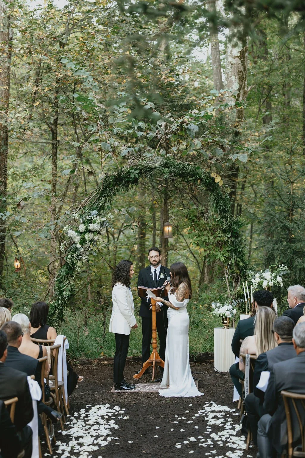 A couple getting married outdoor in a forest, with an officiant at a wooden stand, surrounded by guests seated on wooden chairs, flower arrangements, and a greenery arch.