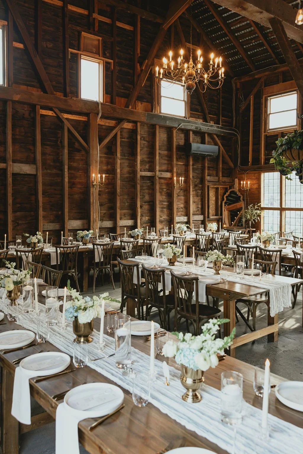 Elegant rustic wedding reception setup with long wooden tables, white floral centerpieces, brass vases, candles, and wood chairs in a barn with high ceilings and chandeliers.