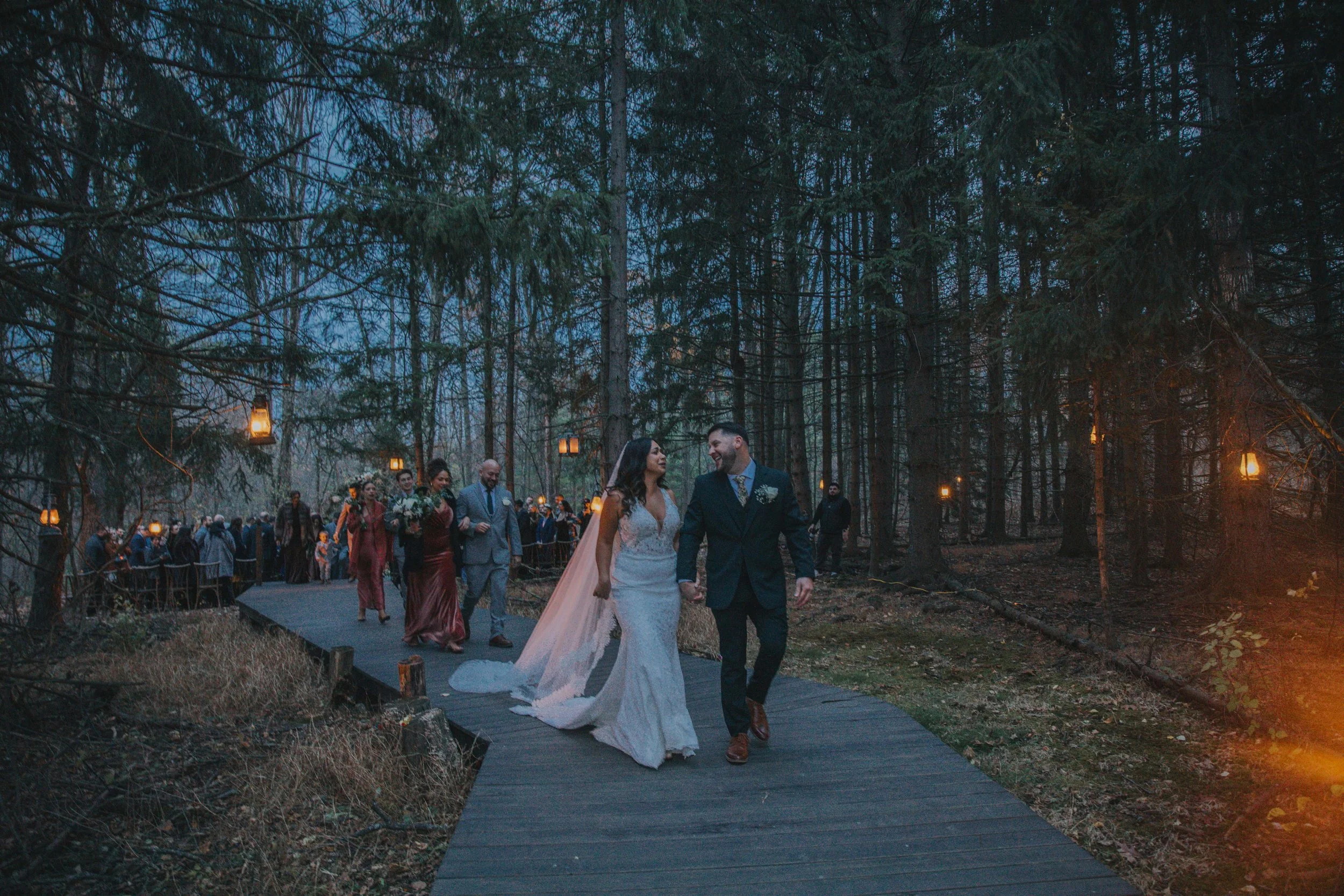 A wedding ceremony outdoors in a forest at dusk, with a bride and groom walking hand in hand on a wooden pathway surrounded by guests and hanging lanterns.