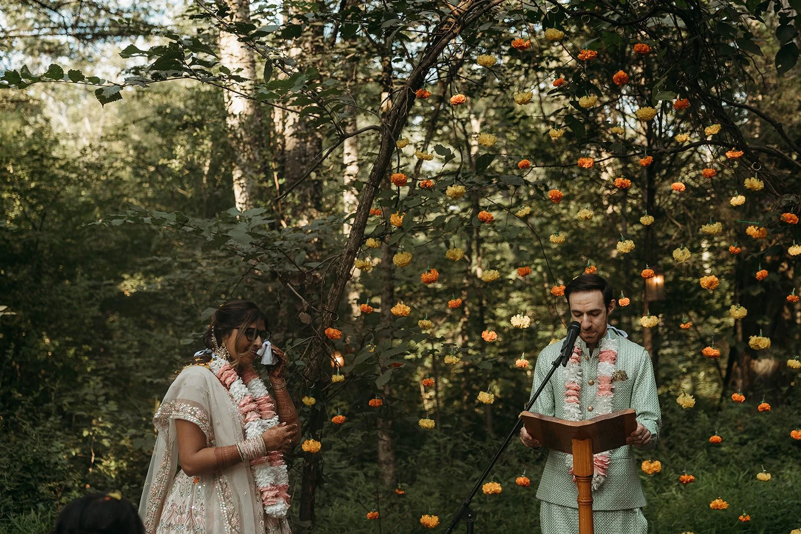A couple in traditional Indian wedding attire participating in a ceremony outdoors, decorated with hanging orange and yellow flowers and surrounded by trees.