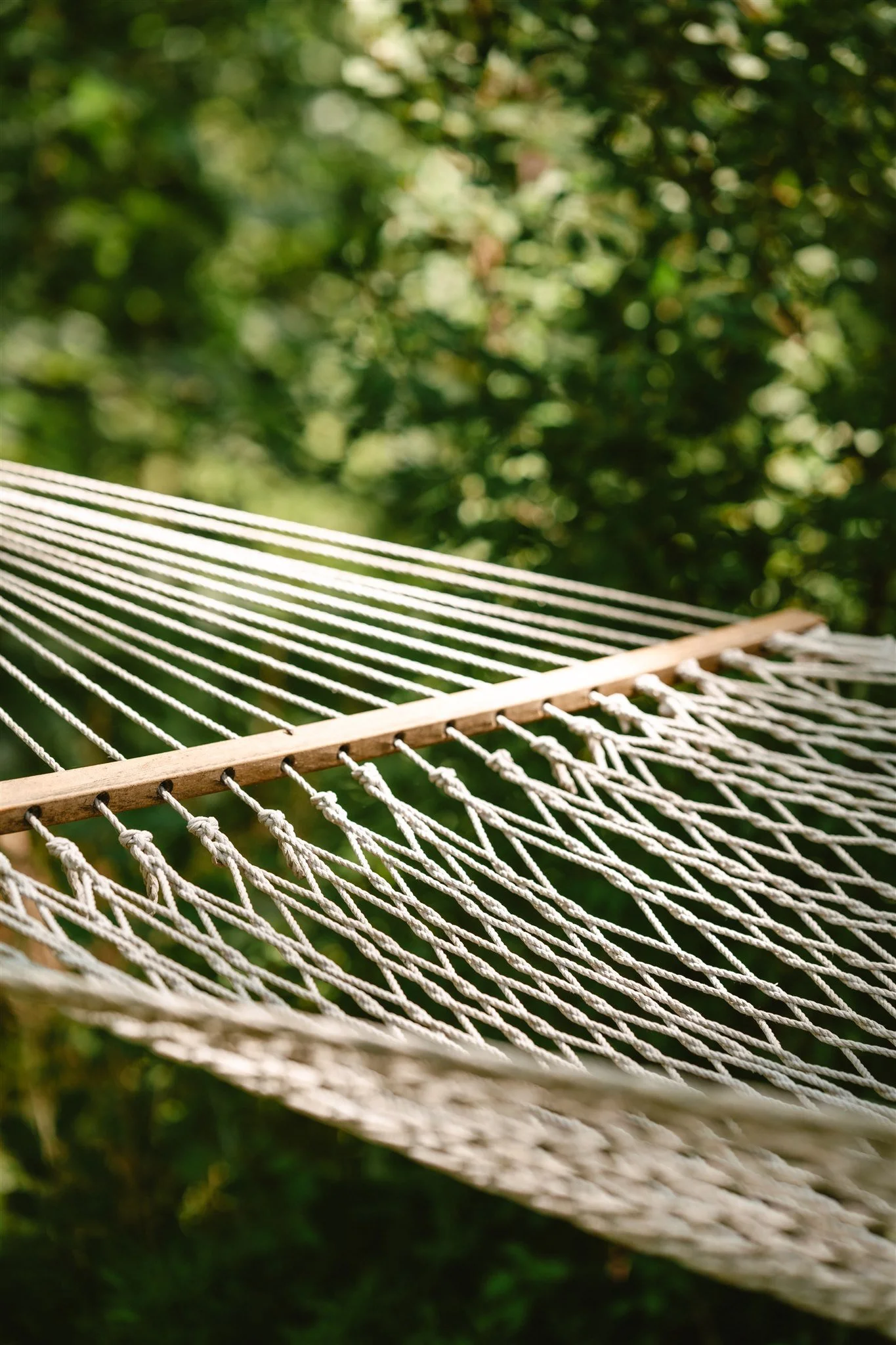 Close-up of a white rope hammock with wooden spreader bars, suspended outdoors with green foliage in the background.