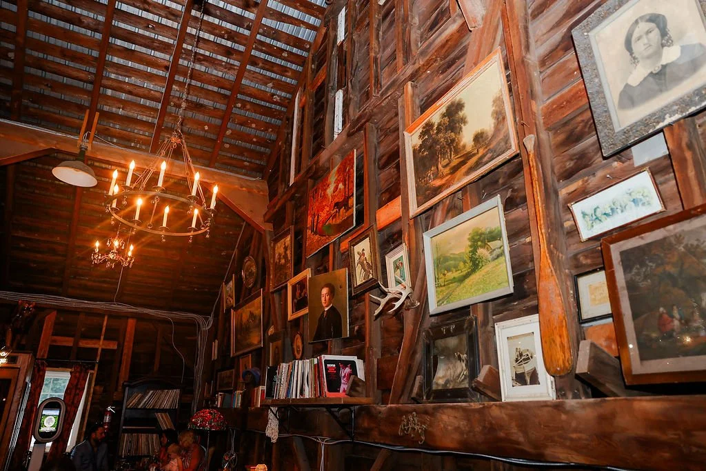 Interior of a rustic wooden room with a high ceiling, decorated with various framed paintings and photographs on the wall. There are chandeliers with lit candles hanging from the ceiling.