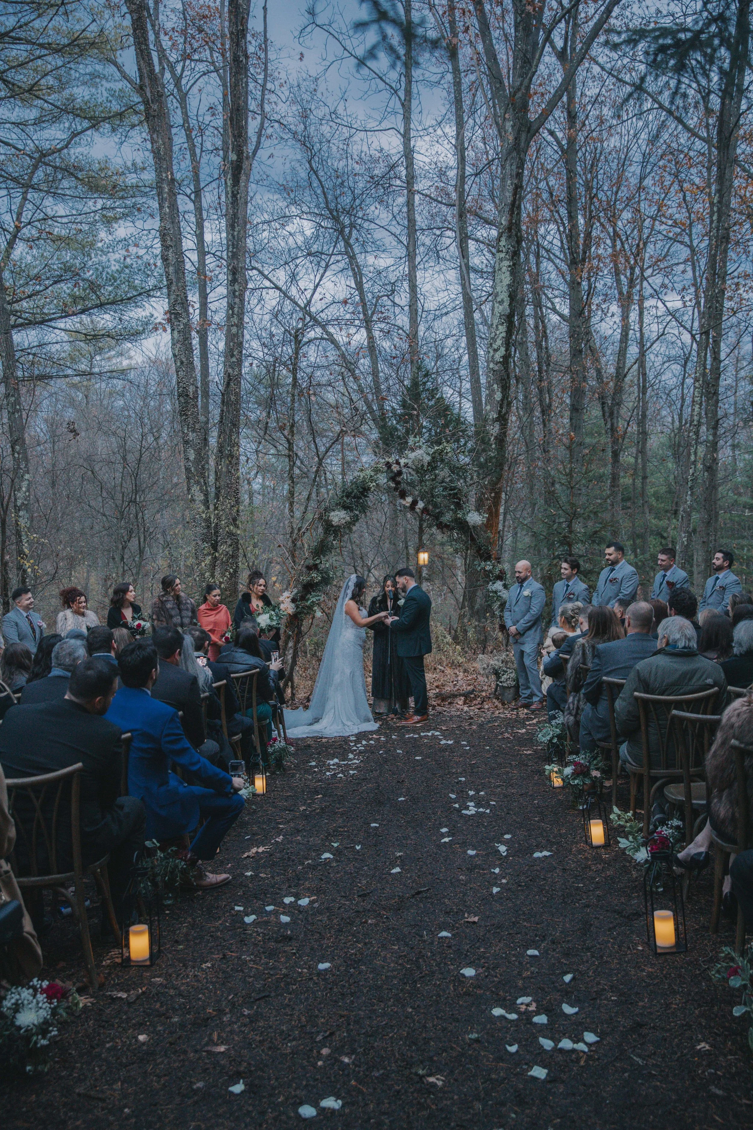 Outdoor wedding ceremony in a wooded area with guests seated on either side, a arch decorated with flowers, and the bride and groom exchanging vows.