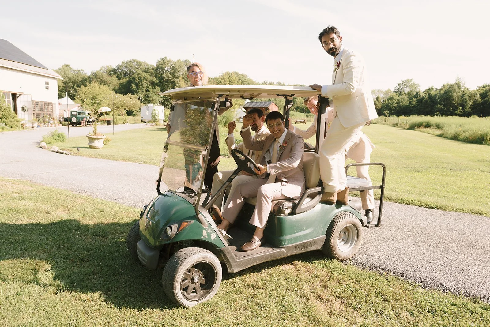 A group of people dressed in formal wedding attire riding on a green golf cart outdoors on a sunny day.