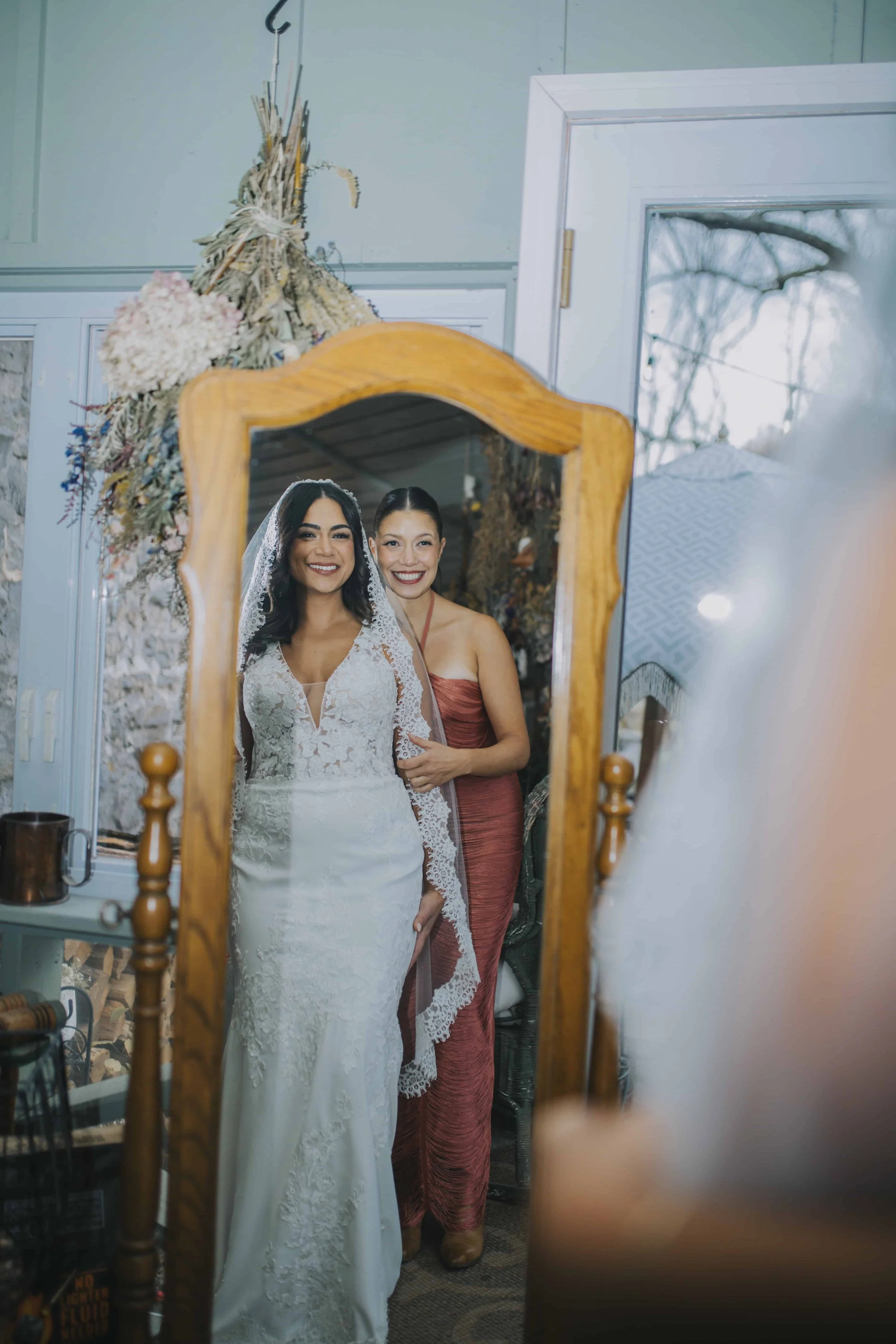 A bride in a white lace wedding dress and veil stands in front of a mirror, smiling, with a woman in a mauve dress beside her, also smiling. The scene is reflected in the mirror, capturing both women.