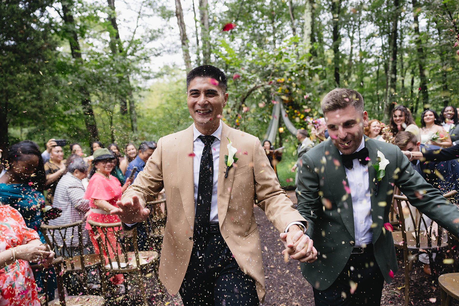 Two men in suits, holding hands and smiling, walking through a colorful outdoor wedding ceremony with guests celebrating and throwing confetti in a forest setting.