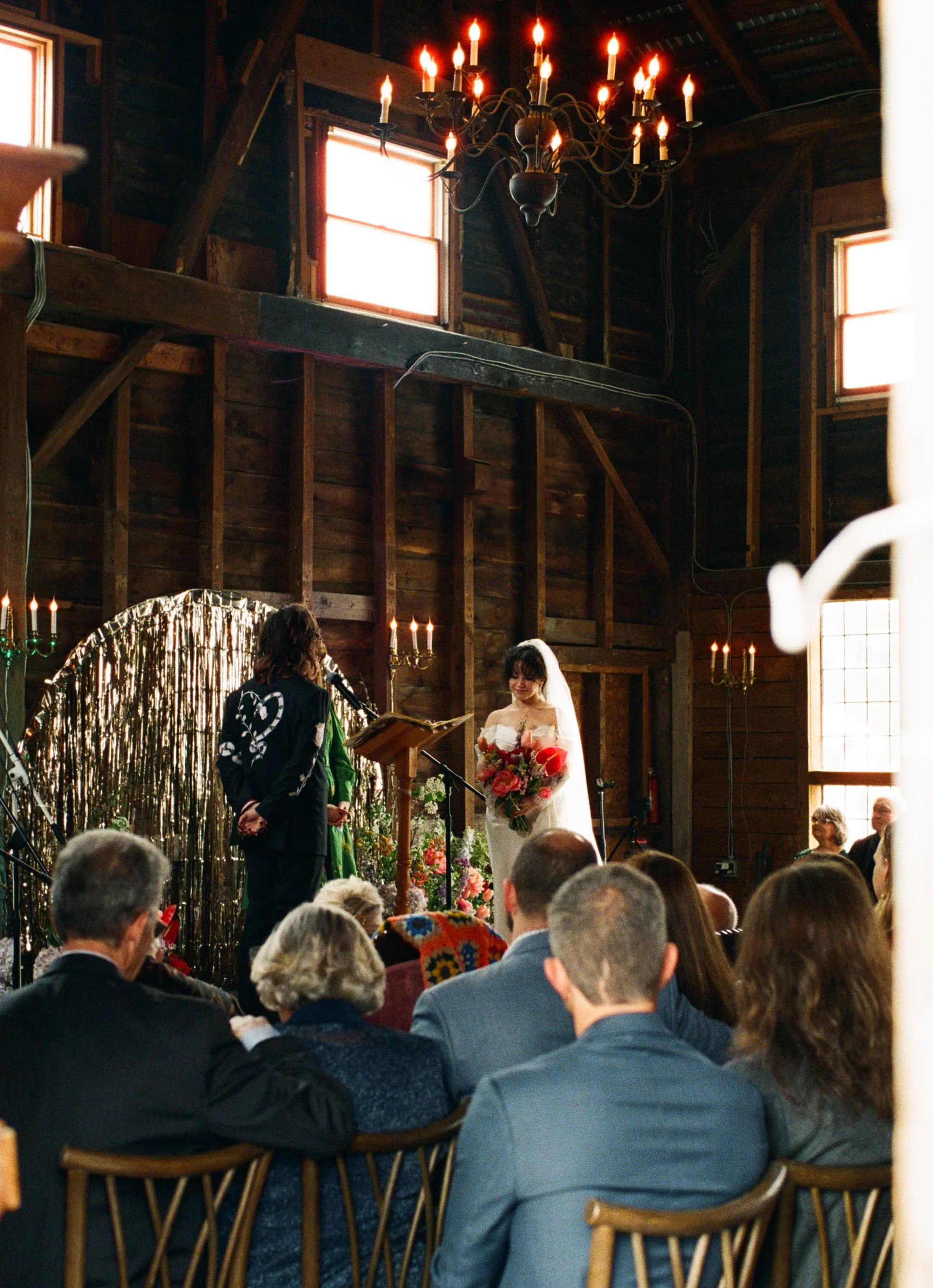 Wedding ceremony inside a rustic wooden barn with a bride holding a bouquet, officiant, and guests seated before a decorated altar.