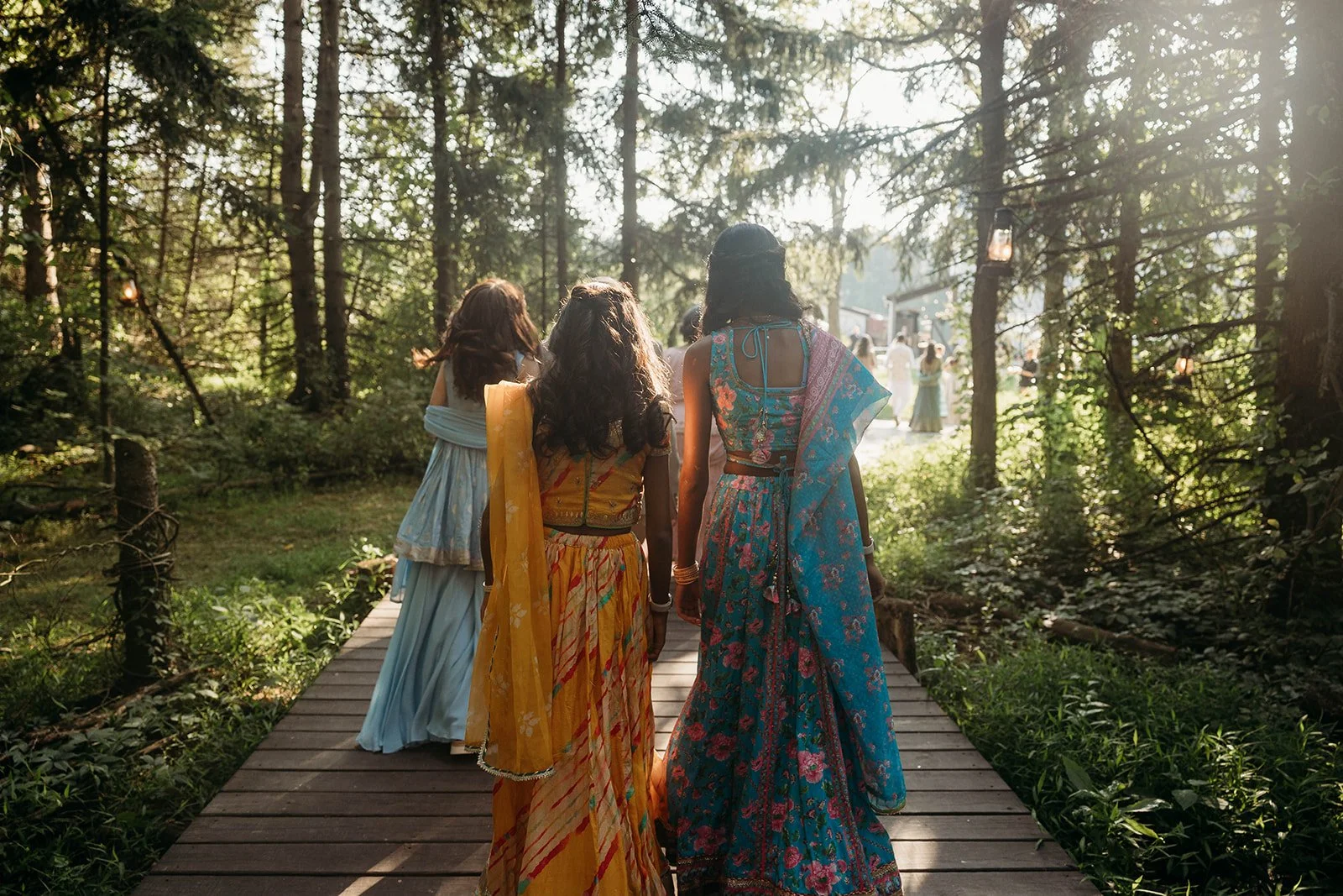 Three women in colorful traditional Indian sarees walking on a wooden pathway through a forested area during a celebration or wedding, with sunlight streaming through the trees.
