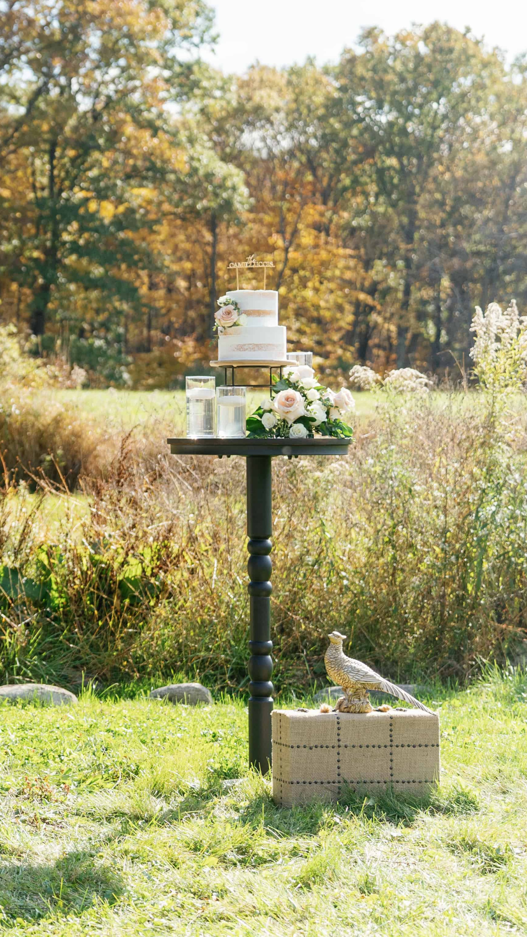 A rustic hightop cocktail table with flowers and a cake, in a woodland setting at Black Walnut Farm