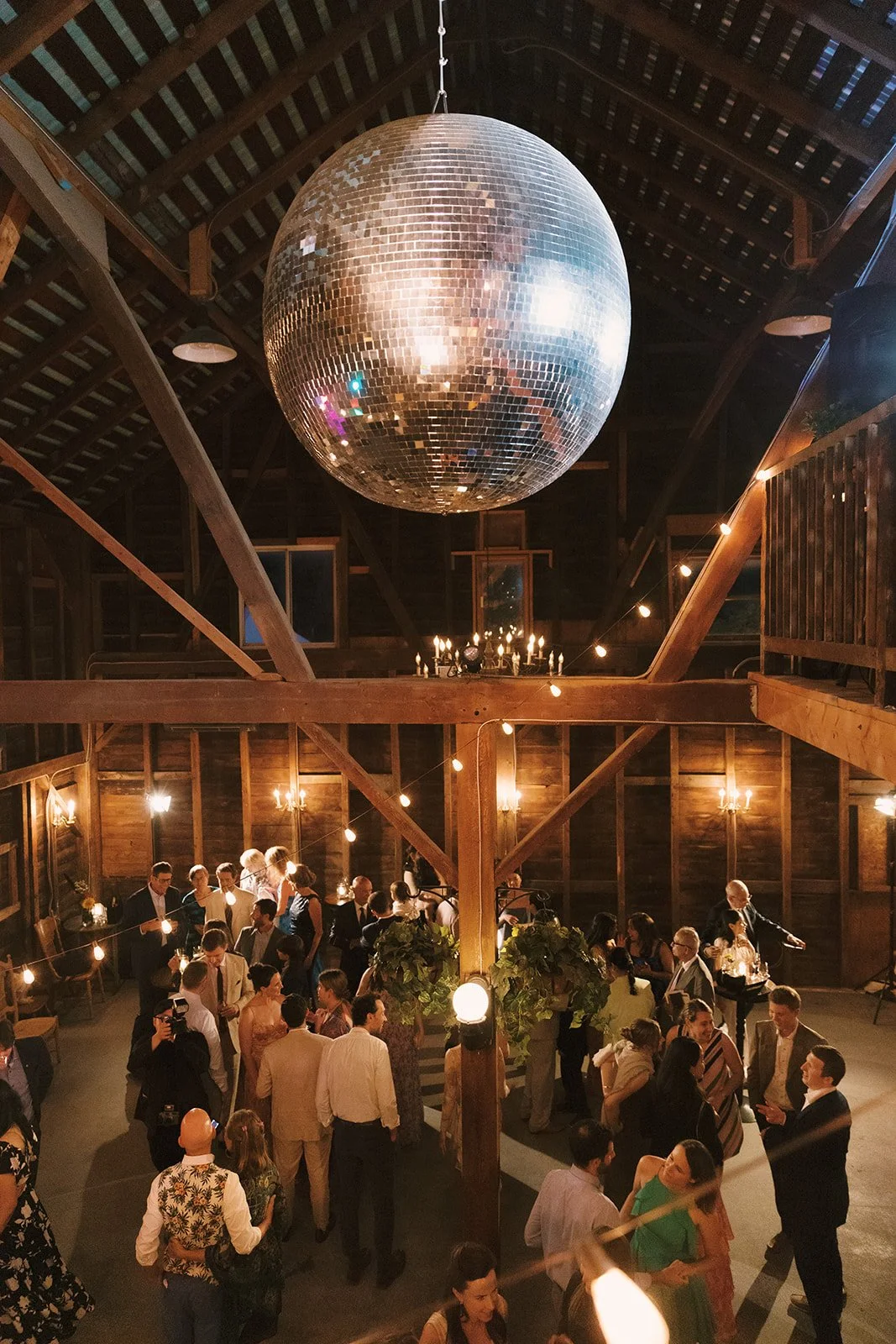A dance party in a wooden barn with a disco ball hanging from the ceiling, guests dancing and socializing, string lights, candles, and plants decorating the space.