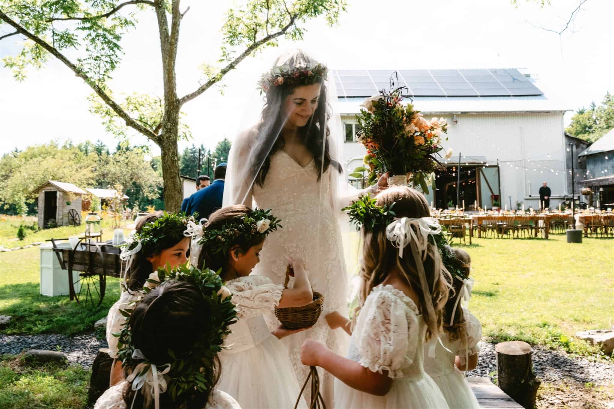 Bride in a white wedding dress with a flower crown and veil, holding a bouquet, surrounded by four young girls with flower crowns and dresses, in a garden setting with trees, a barn, and outdoor wedding decorations.