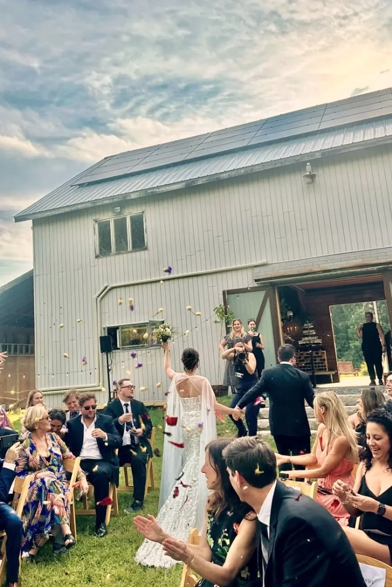 A wedding celebration outdoors with guests seated in chairs, a bride and groom holding hands, and a photographer taking pictures, with a barn-style building in the background and a partly cloudy sky overhead.