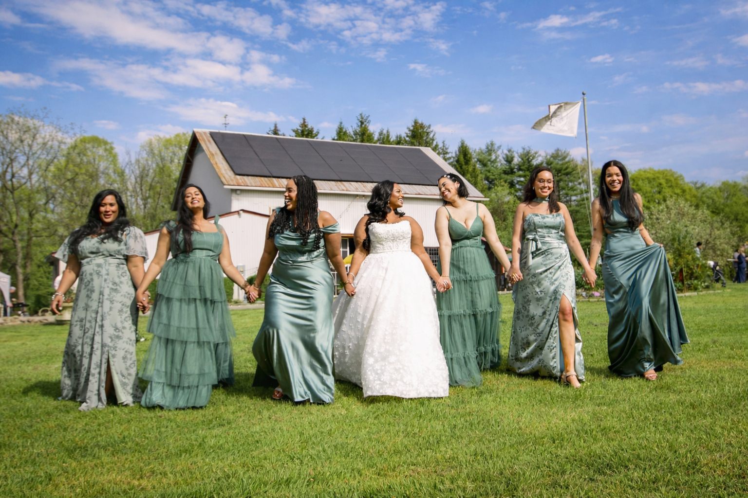 A group of women in formal dresses holding hands and walking outdoors on a sunny day, with a barn, trees, and a flag in the background.