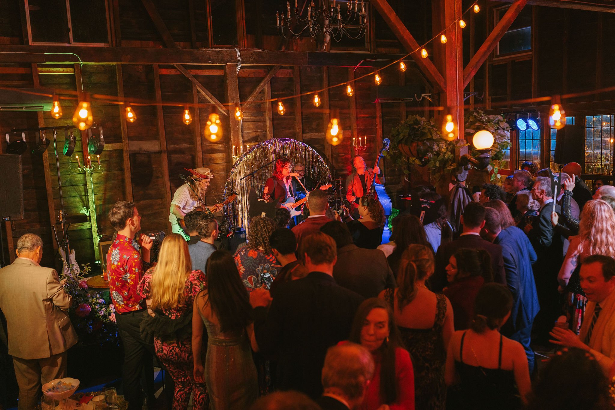 A lively indoor concert with a band performing on stage and an audience dancing and enjoying the music. The venue has wooden walls, warm lighting, and string lights hanging from the ceiling.