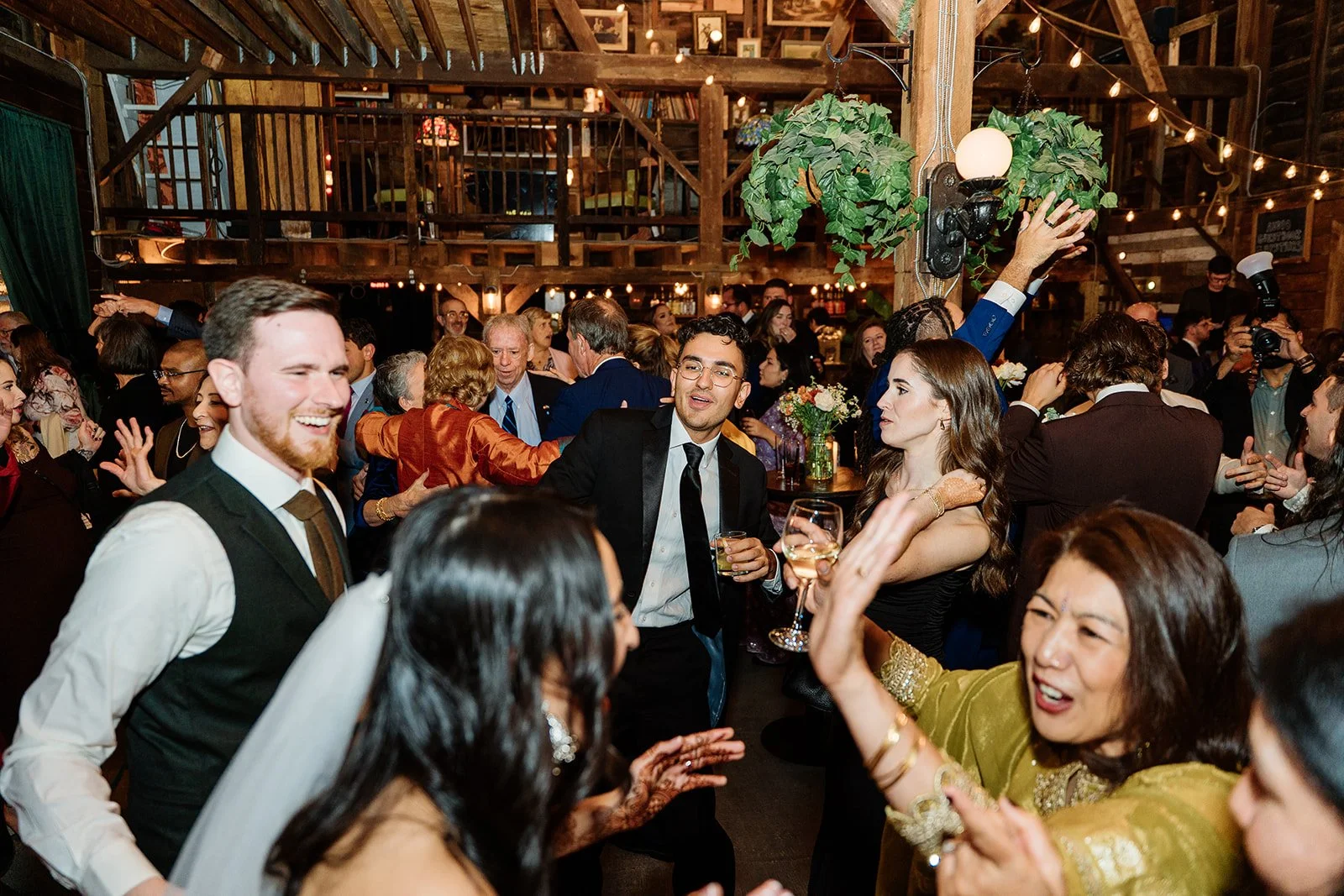People dancing and enjoying a celebration in a rustic, wooden barn with string lights, floral arrangements, and a lively atmosphere.