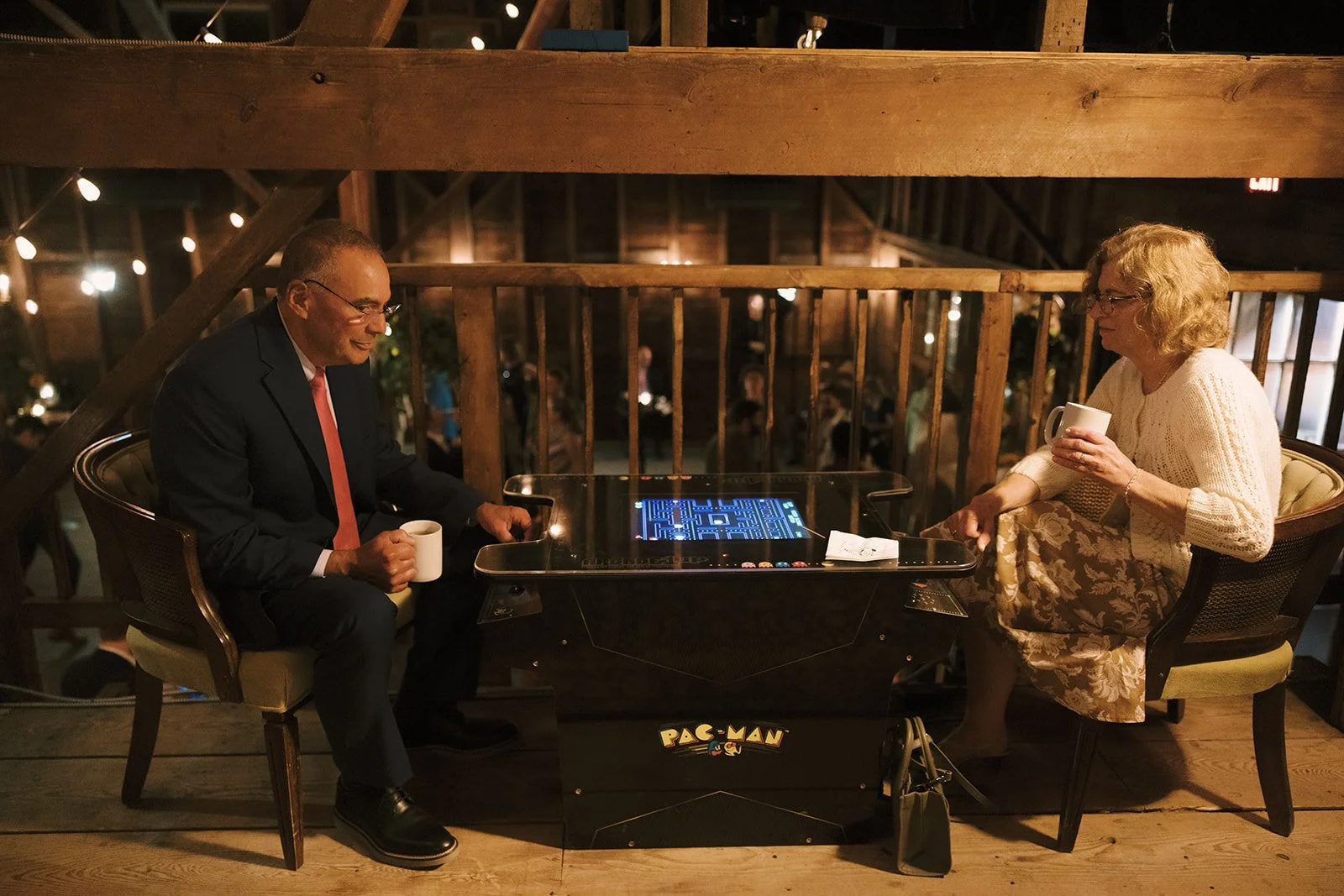A man and woman sitting across from each other at a Pac-Man arcade game table, each holding a mug, in a warmly lit wooden-finished indoor setting, possibly a restaurant or lounge.