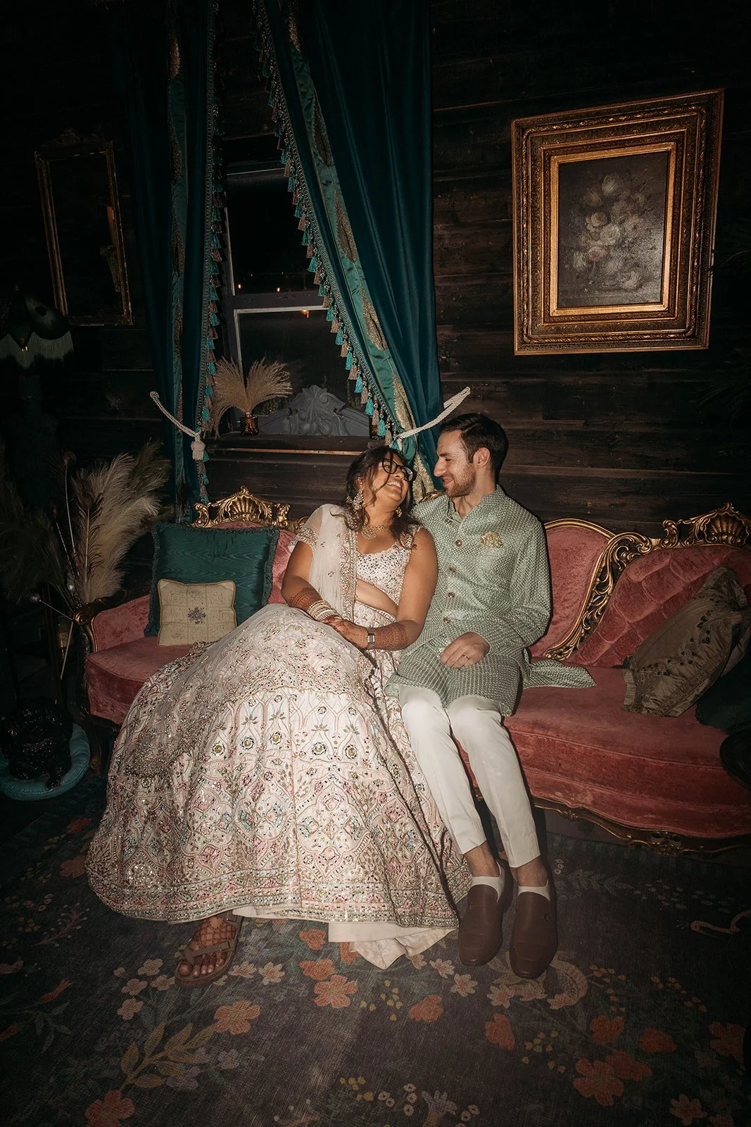 A couple dressed in traditional Indian attire sitting on a vintage pink sofa with ornate gold details, sharing a joyful moment indoors with dark wooden walls, framed artwork, and tall teal curtains.