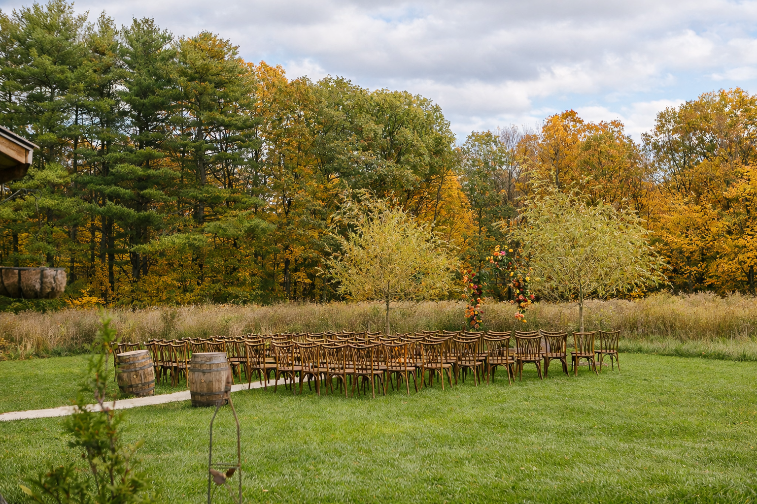 Outdoor setting with a grassy lawn, a line of wooden chairs, and trees with fall foliage, a large arch decorated with flowers, and a cloudy sky.