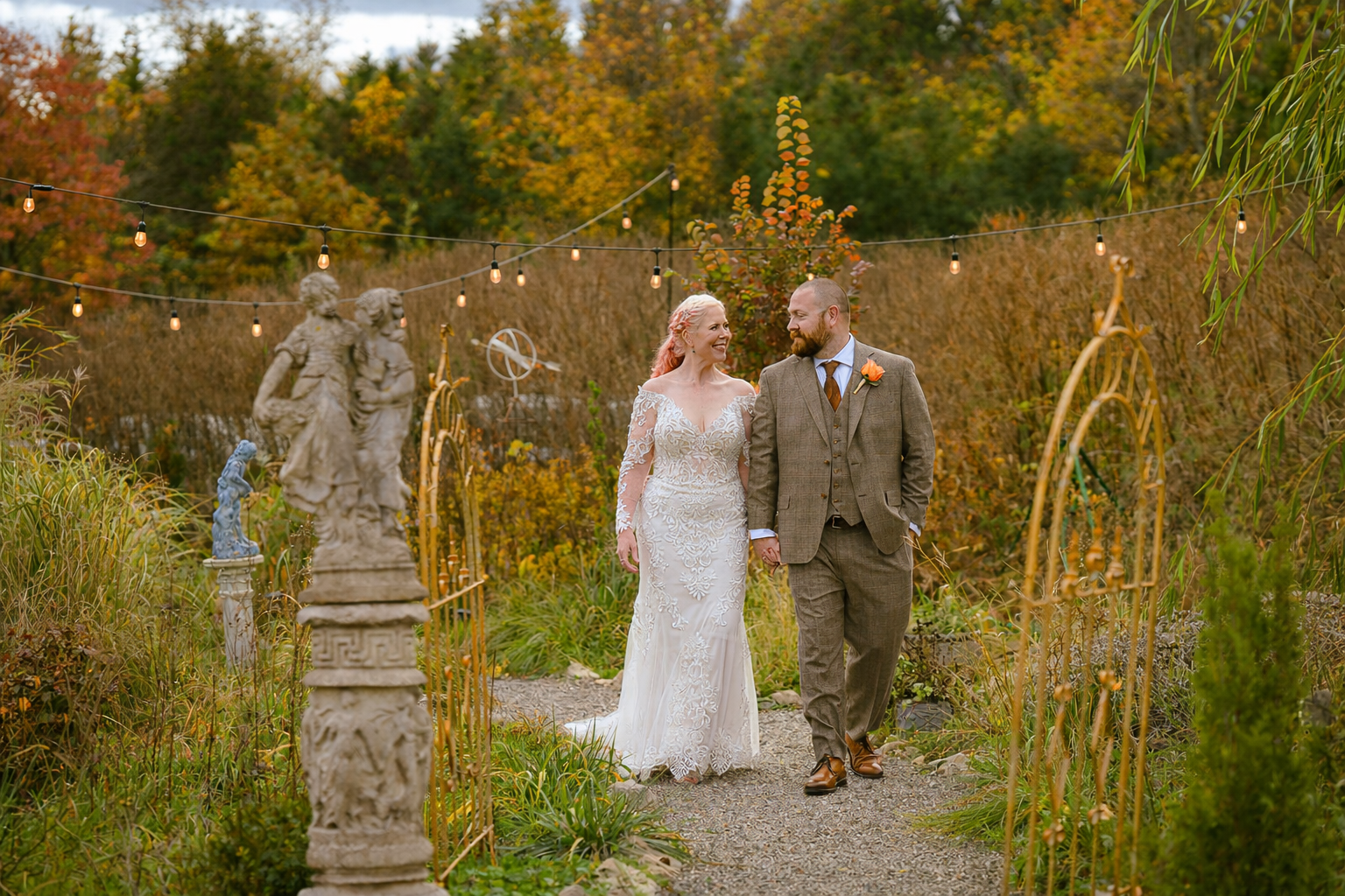 A bride and groom walking hand in hand in a garden during autumn, surrounded by statues and string lights, smiling at each other.