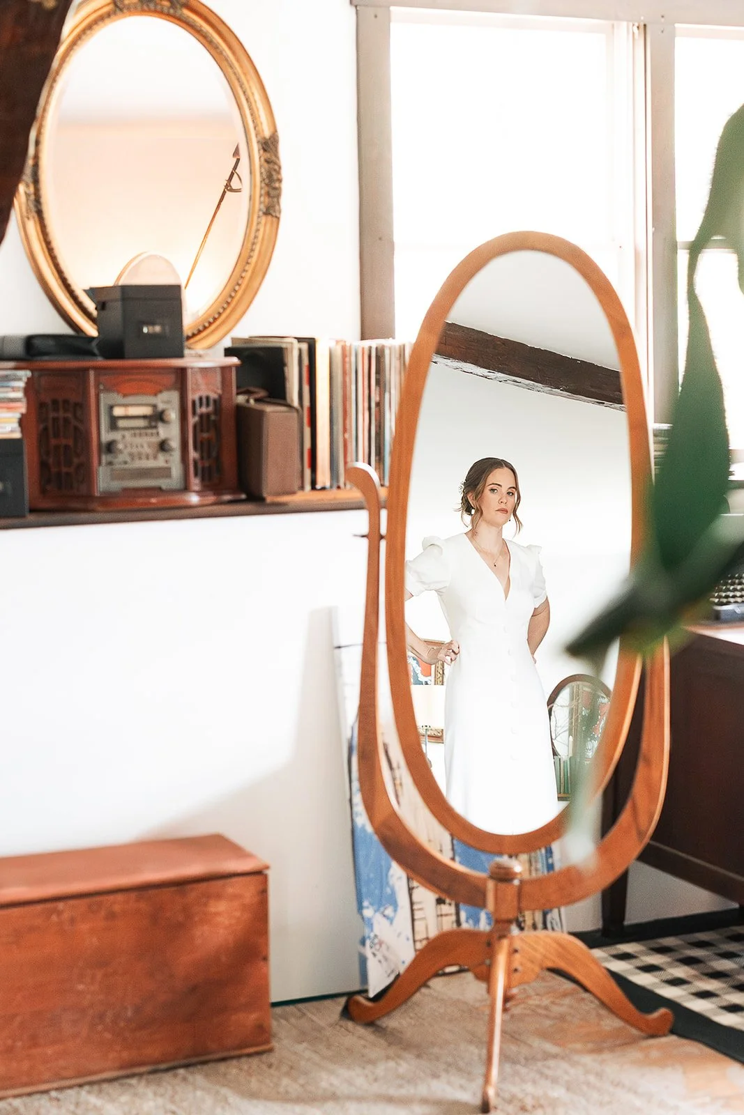 A woman dressed in a white dress is seen through a standing mirror with a wooden frame in a room with vintage decor. The mirror reflects her pose with hands on her hips.