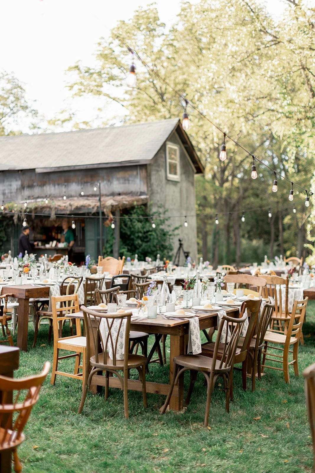 Outdoor rustic wedding reception setup with wooden tables and chairs, floral centerpieces, string lights, and a barn in the background, surrounded by trees.
