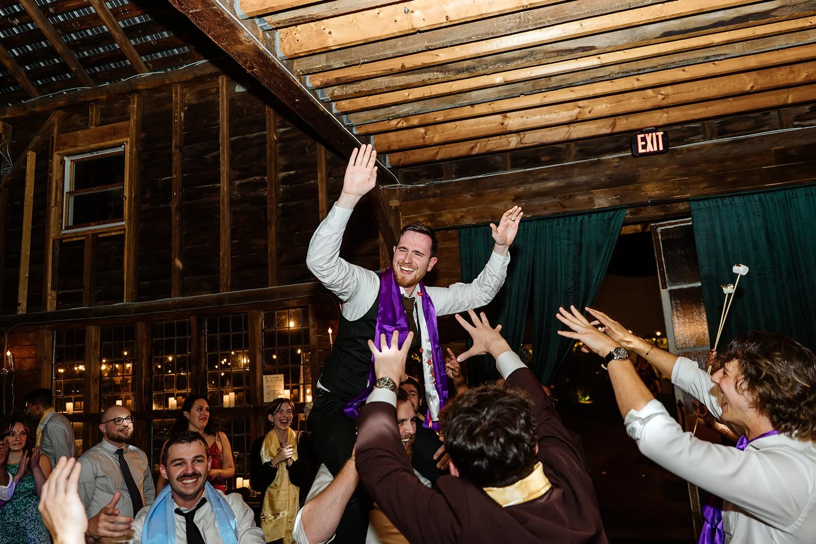 People celebrating at a party, with a man being lifted on others' shoulders, smiling and raising his hands, in a wooden venue with candles and decorations.