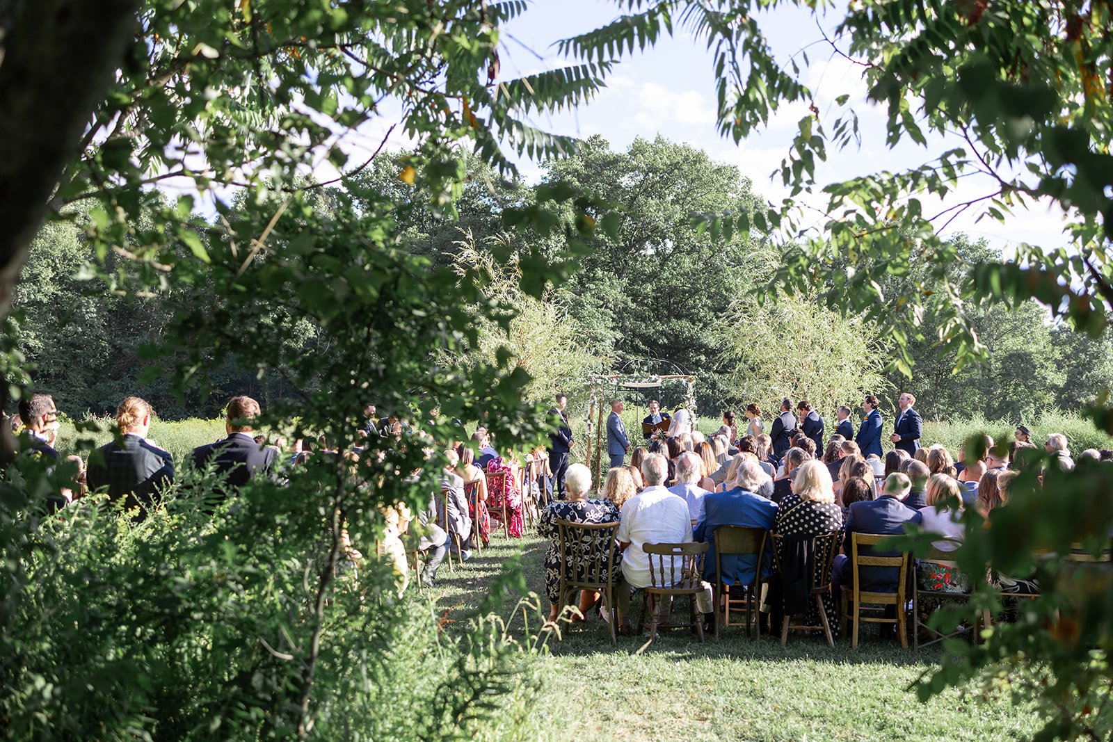 Outdoor wedding ceremony with guests seated on wooden chairs, a bride and groom standing under a rustic arch, surrounded by trees and greenery, on a sunny day.