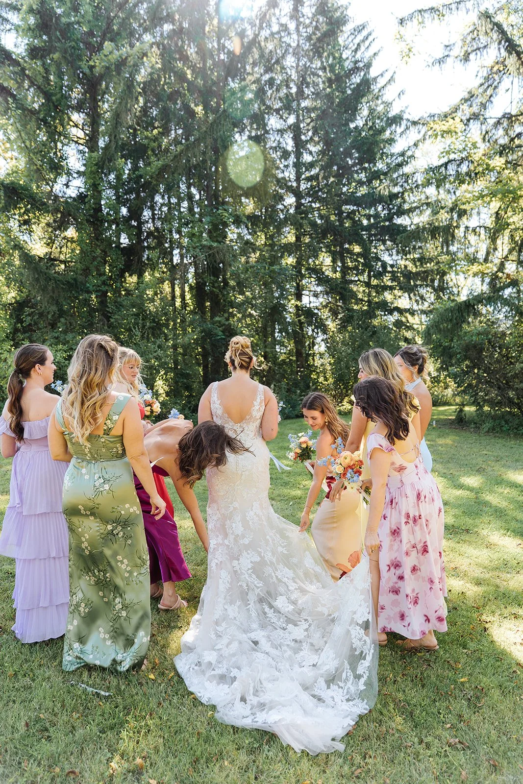 A bride in a white lace wedding dress standing outdoors with her bridesmaids and friends in colorful dresses, surrounded by tall trees and sunlight.
