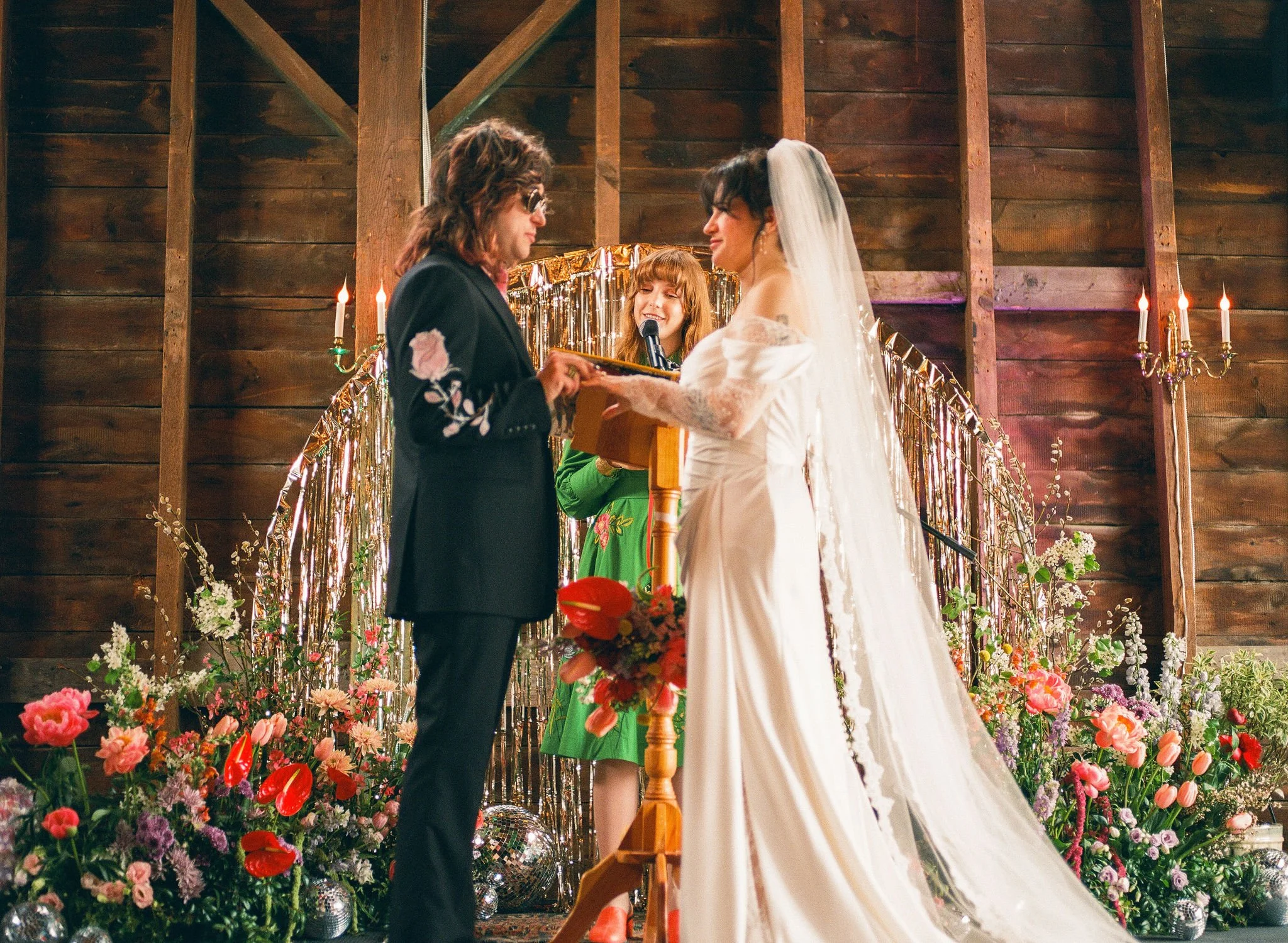 A bride and groom stand at an altar of gold tinsel and exotic flowers in the Barn at Black Walnut Farm