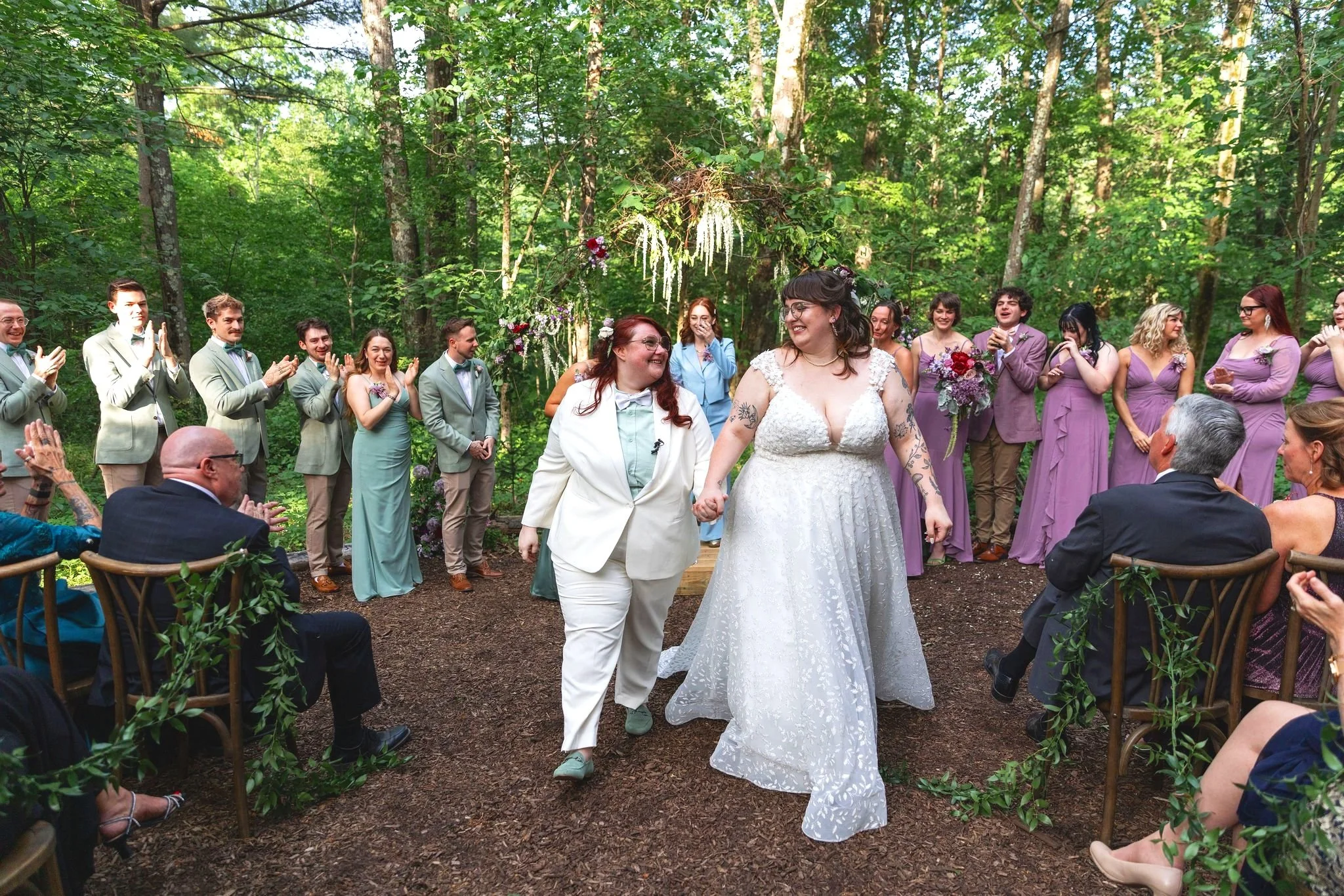 A wedding ceremony takes place in a forest, with two brides holding hands and walking down an aisle. One bride is wearing a white dress with floral lace details, and the other is wearing a white suit. Guests are seated and standing around, clapping a