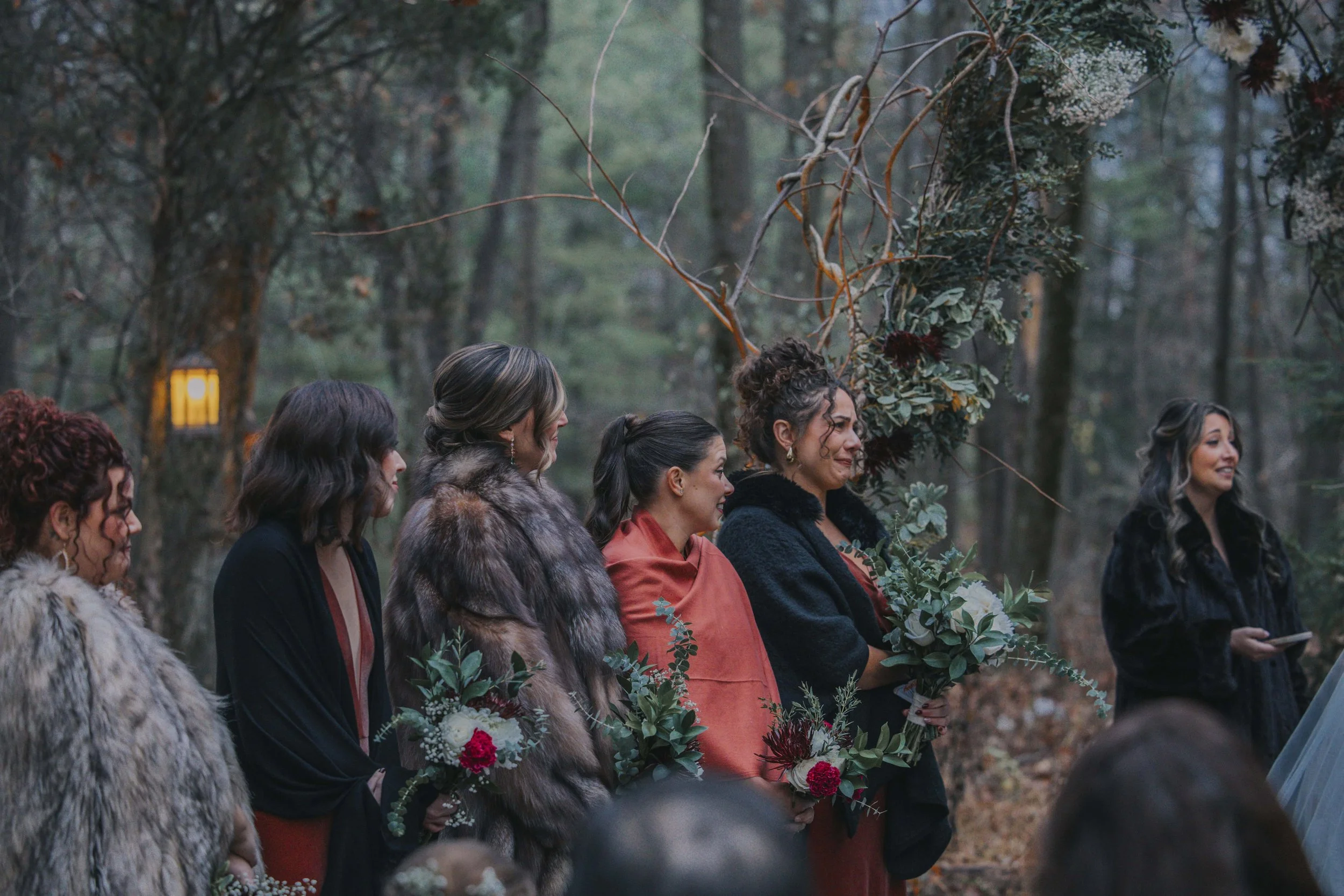 A group of women in formal attire standing in a forest, participating in a wedding ceremony, holding bouquets of flowers.