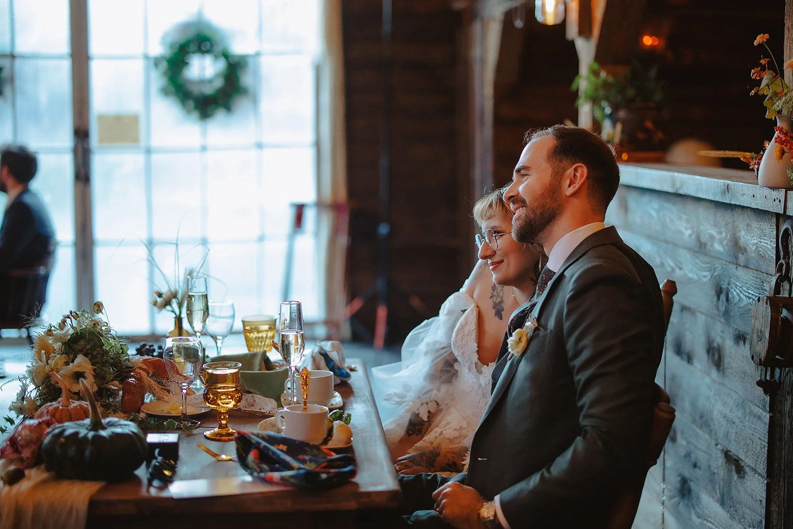 A newlywed couple sits side by side at a wedding reception table, smiling. The bride has short blonde hair, glasses, and tattoos, wearing a white dress. The groom has a beard and mustache, wearing a dark suit with a boutonnière. The table is decorate