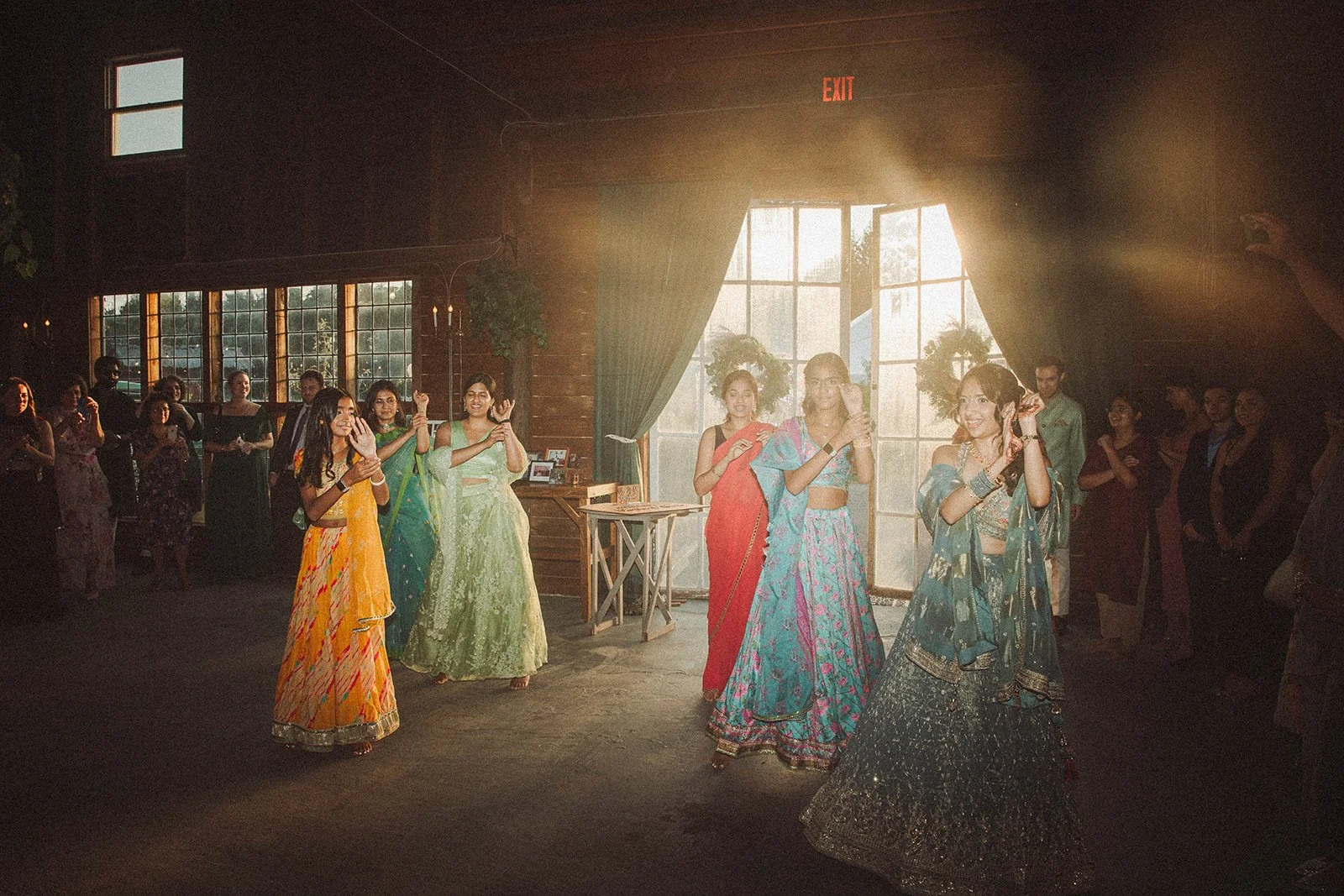 A group of women in colorful traditional Indian attire dancing inside a rustic indoor space with large windows and green curtains, during a celebration or wedding event.