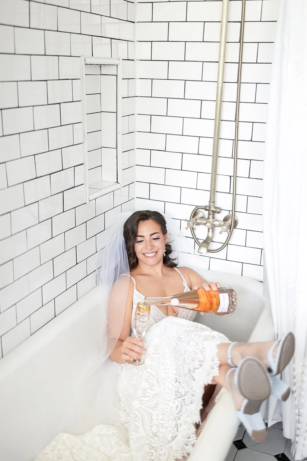 A woman dressed as a bride with a veil, sitting in a clawfoot bathtub, smiling, holding a bottle of liquor, with her feet resting on the edge of the tub.