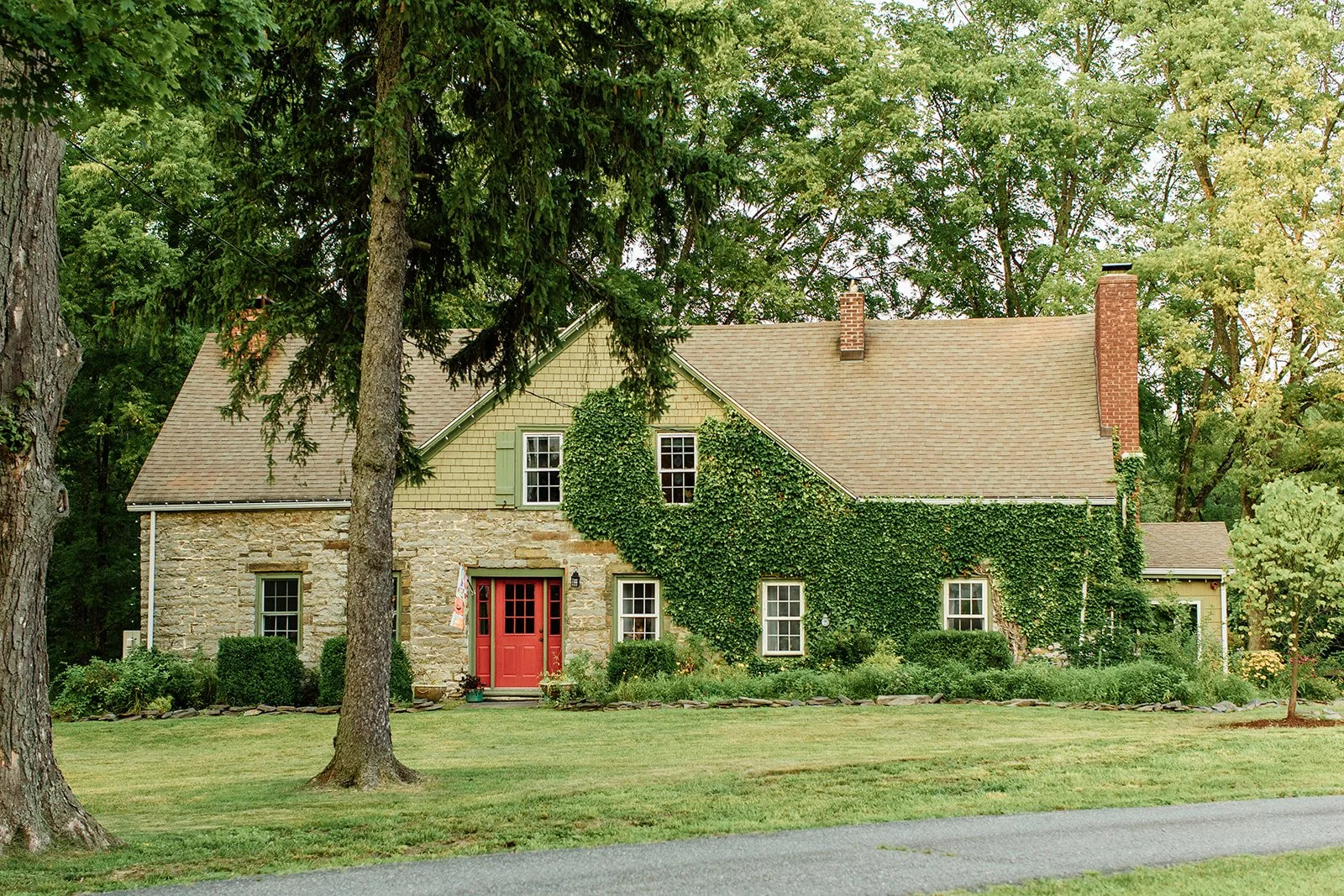 A charming house with stone and yellow siding exterior, surrounded by lush greenery and tall trees, with a well-maintained lawn and a red front door.