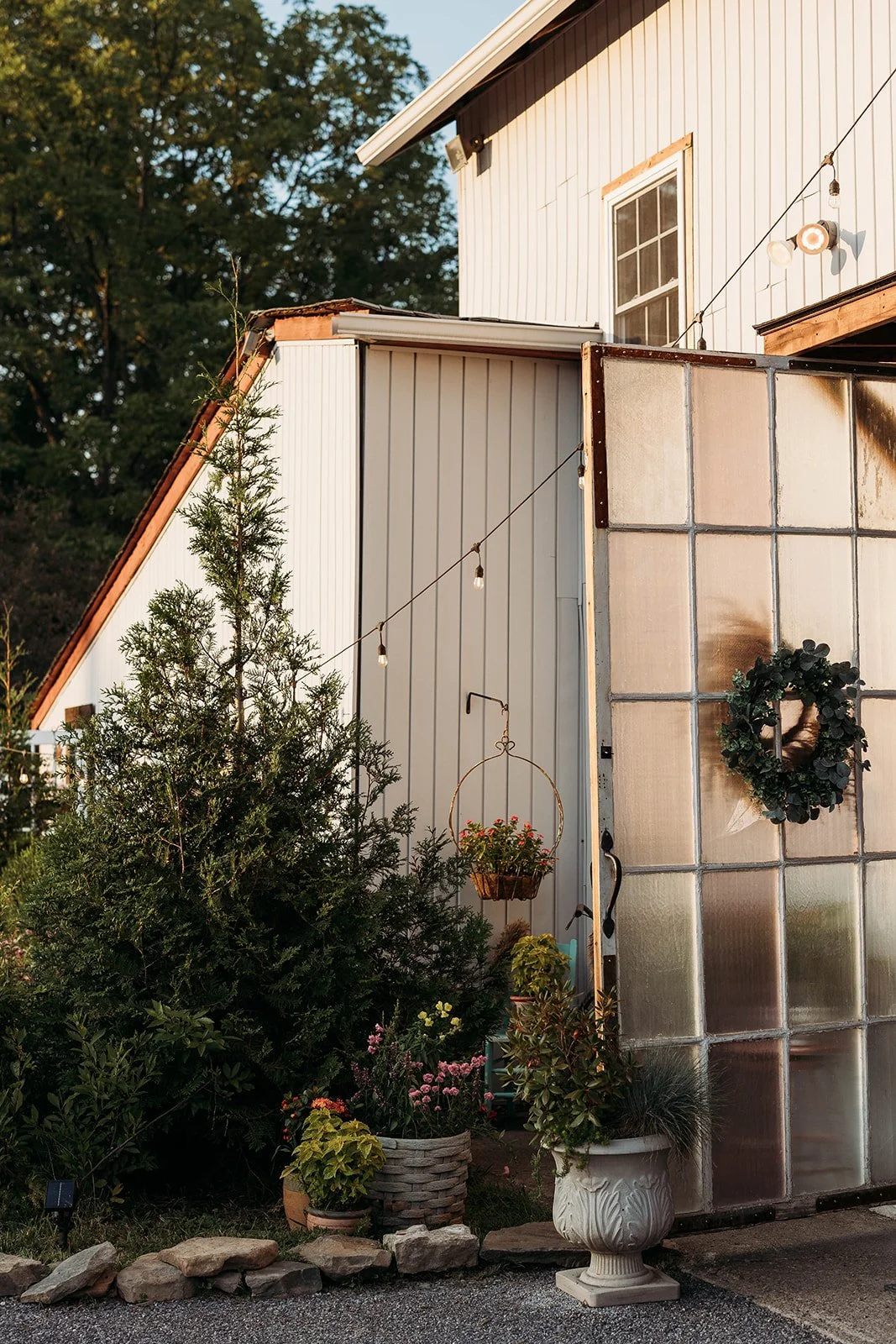 Exterior of a house with a greenhouse attached, decorated with a wreath, potted plants, hanging flower basket, and string lights in the evening.