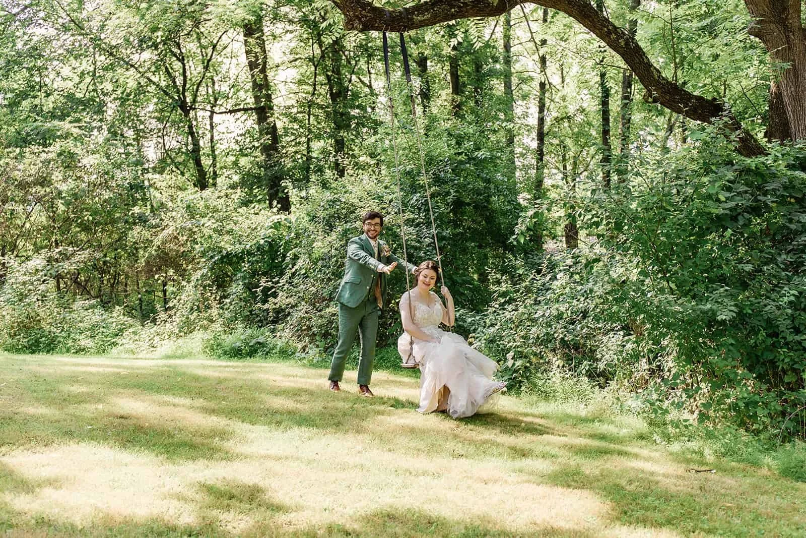 Bride and groom, with the groom pushing the bride on a swing in a lush, green wooded area during daylight.