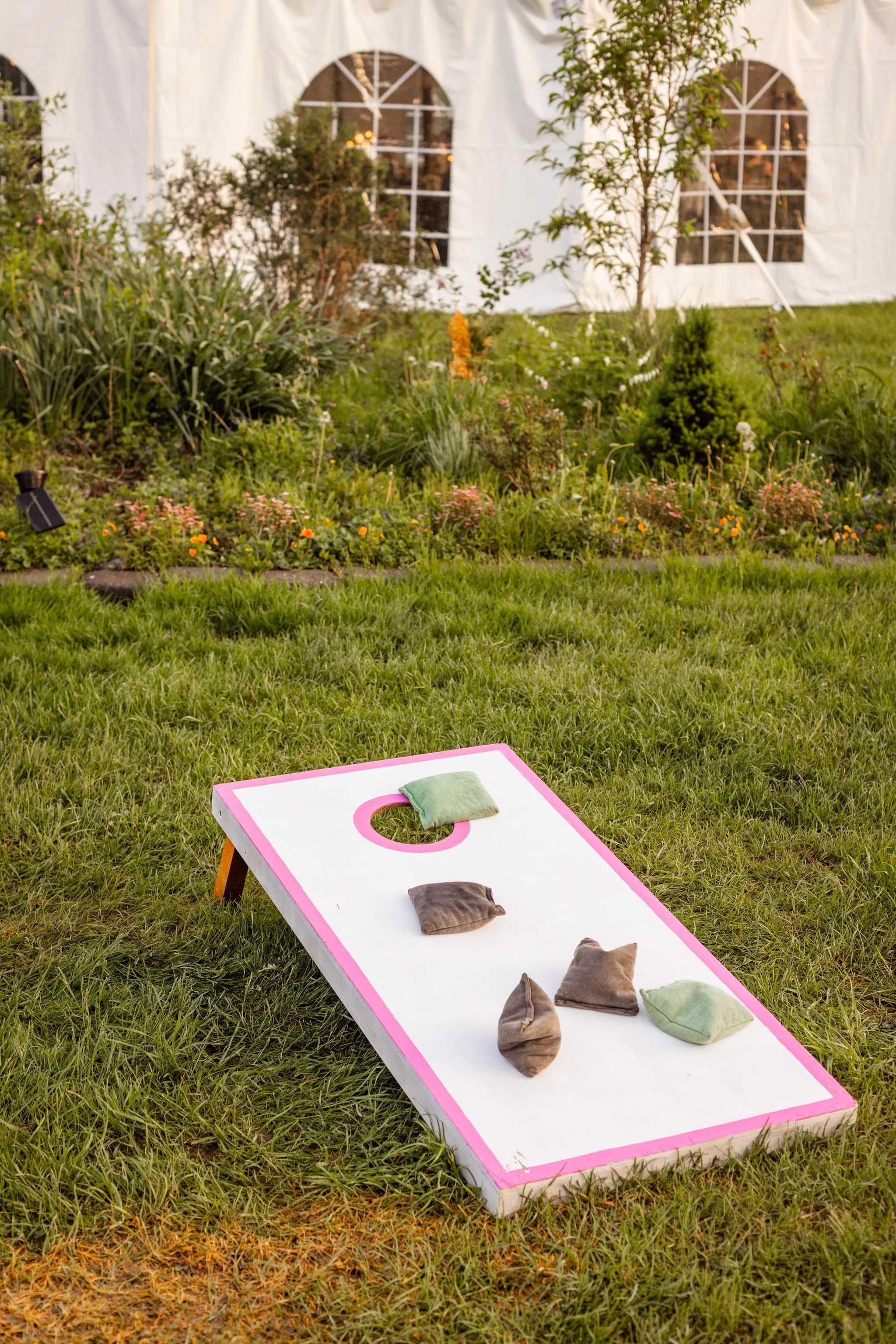 A cornhole game with a white and pink board set up on grass, with four bags placed on it, in front of a garden with plants and a white tent with arched windows.