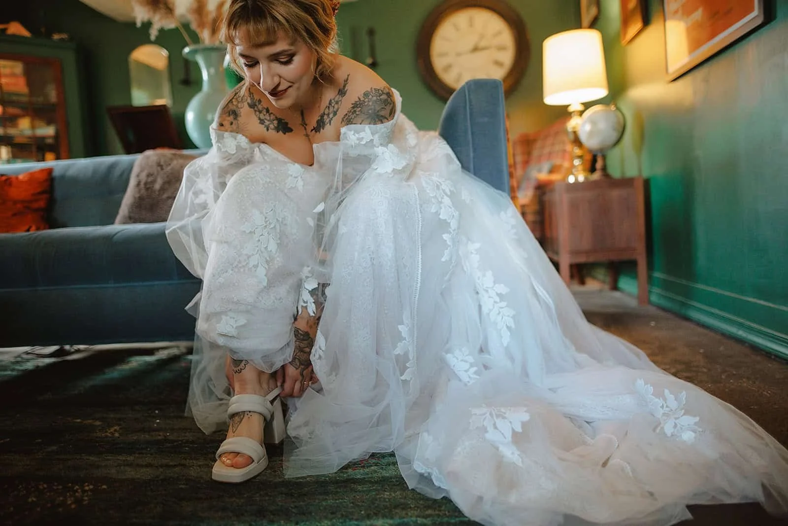 A bride in a gown exposing many tattoos buckles her wedding shoe at Black Walnut Farm
