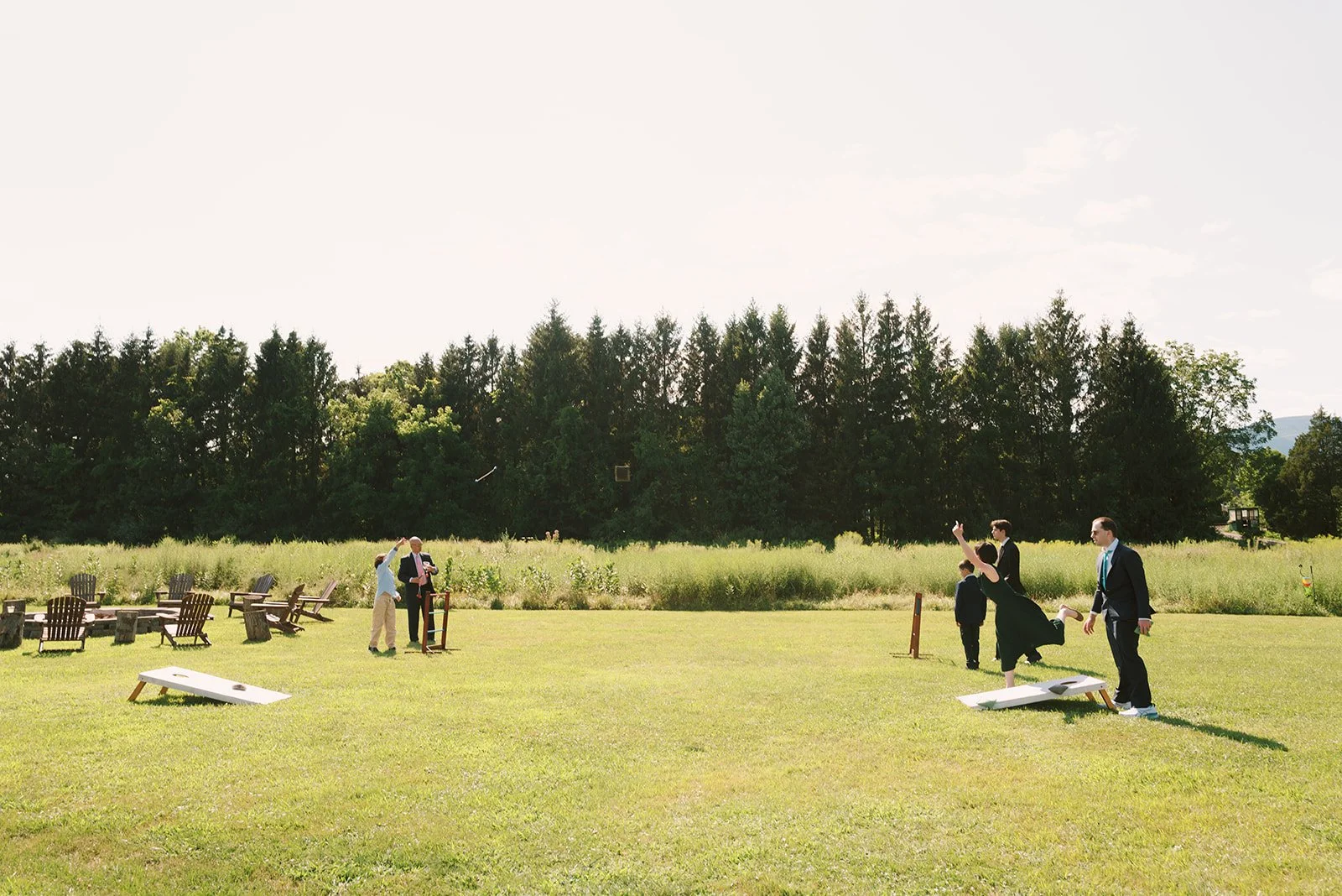 People playing cornhole and enjoying an outdoor gathering on a grassy field with trees in the background.