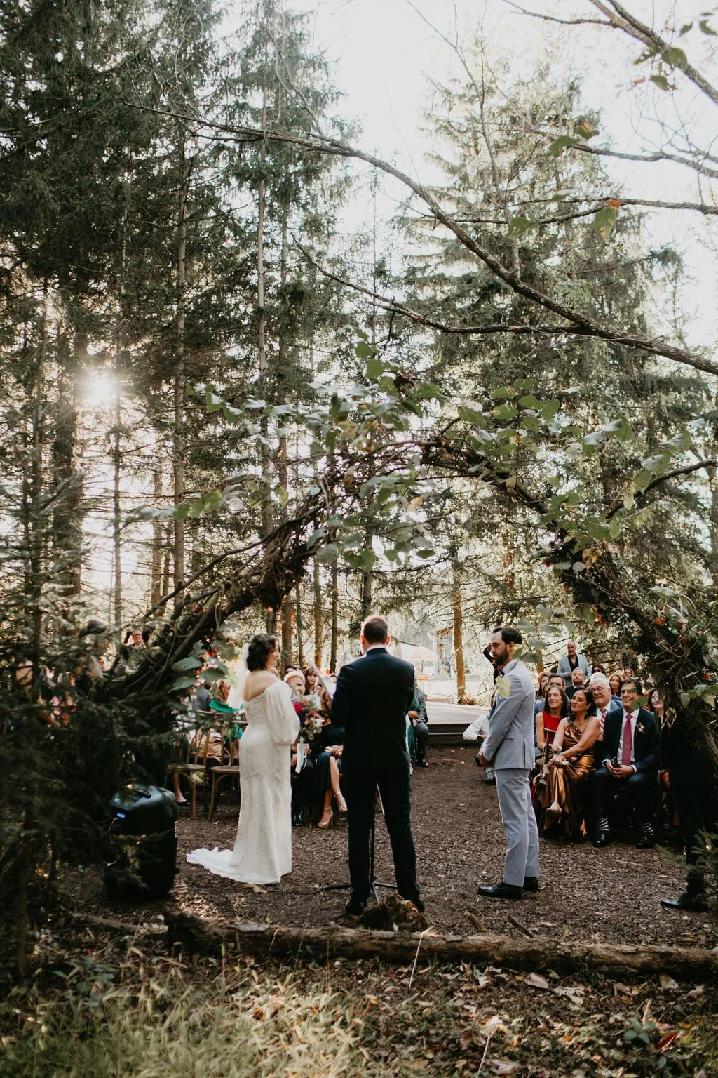 A wedding ceremony taking place outdoors in a wooded area, with guests seated on either side of the couple and officiant under a natural arch made of branches and greenery.