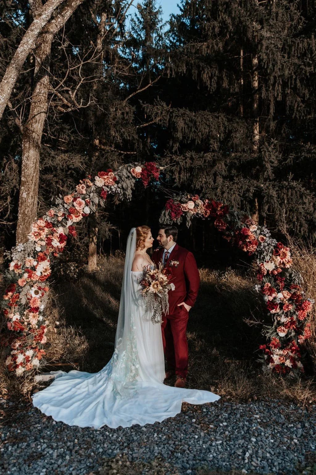 A bride and groom stand under a floral arch in a wooded outdoor setting during dusk.